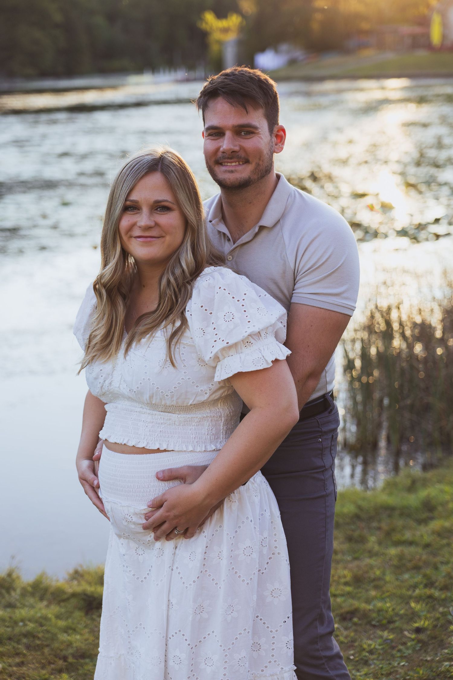 Couple posing outdoors near a lake at golden hour, pregnant woman in white dress with partner embracing her bump.