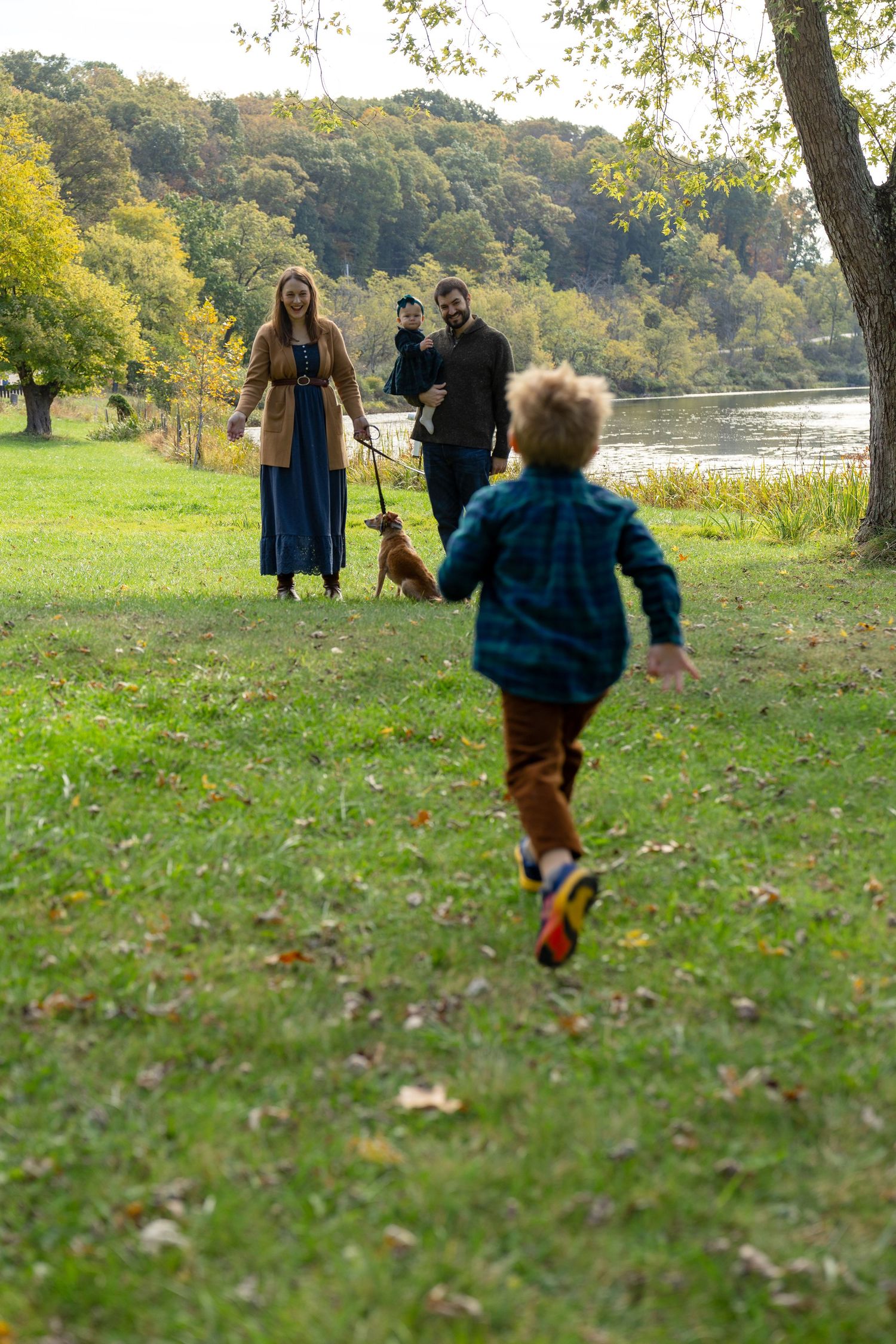 Family enjoying autumn park walk by river, child running toward parents holding baby and dog on leash.