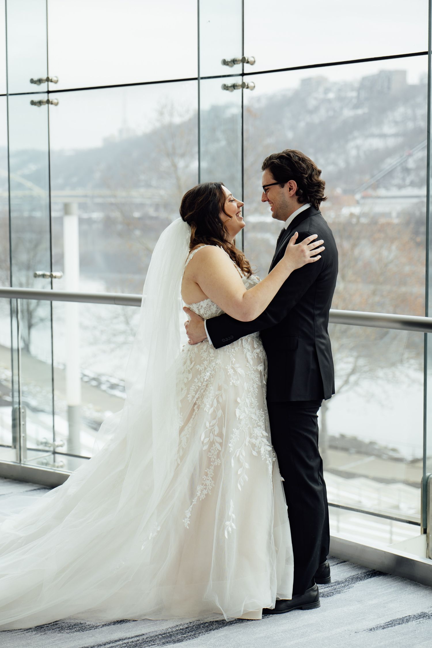 Bride and groom embrace in front of large windows with snowy winter landscape, bride wearing lace gown with veil.