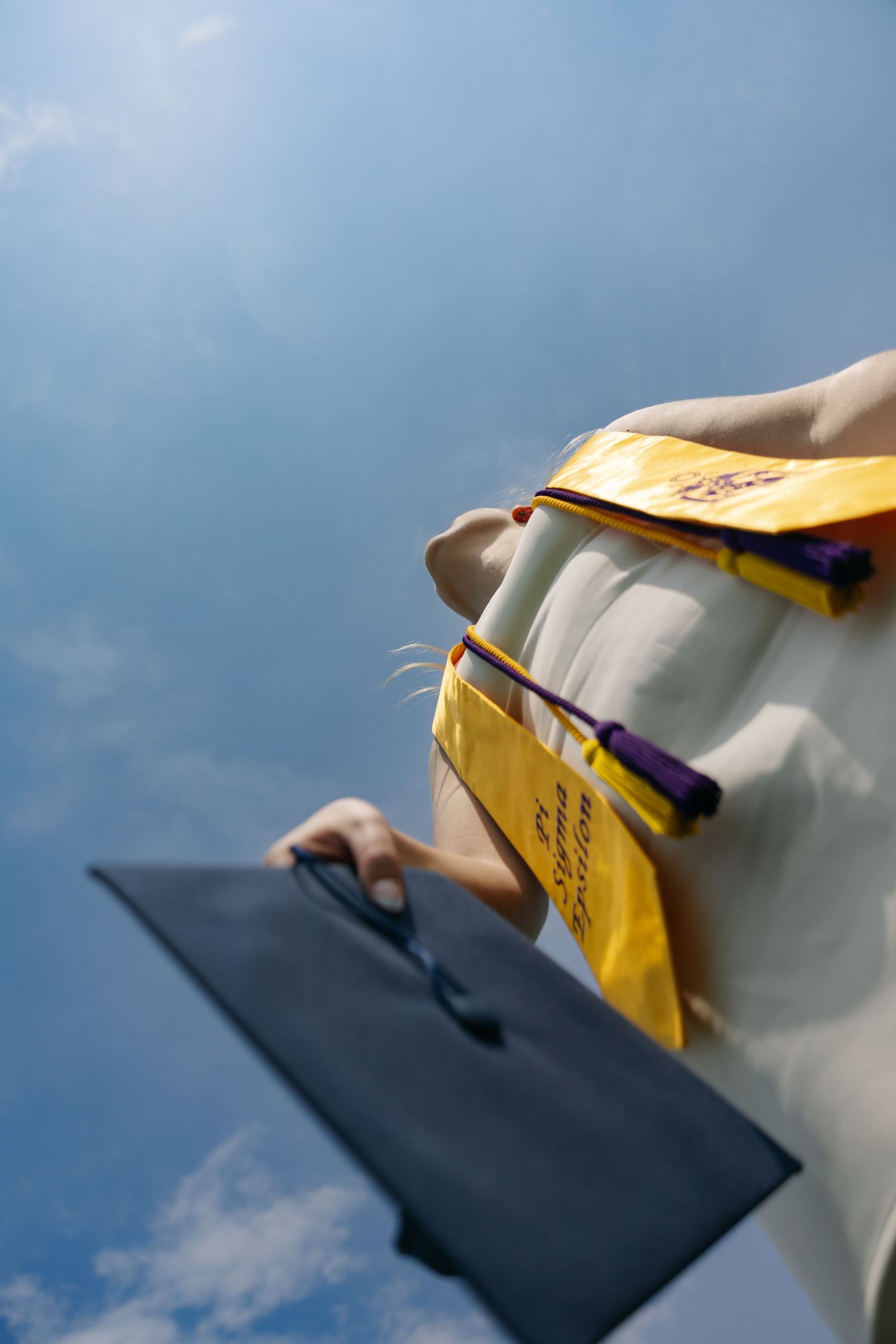 Graduate holding cap against blue sky, wearing white gown with yellow and purple academic regalia at graduation ceremony.