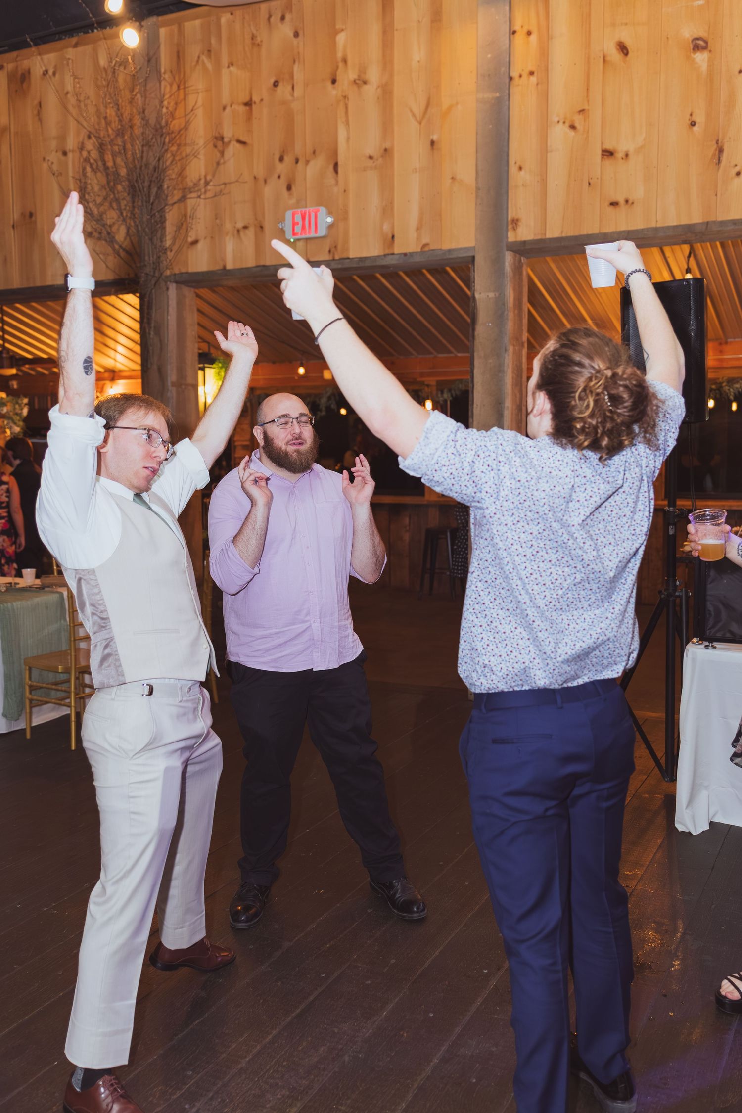 Three men dancing energetically at a rustic barn wedding reception, two with arms raised high.
