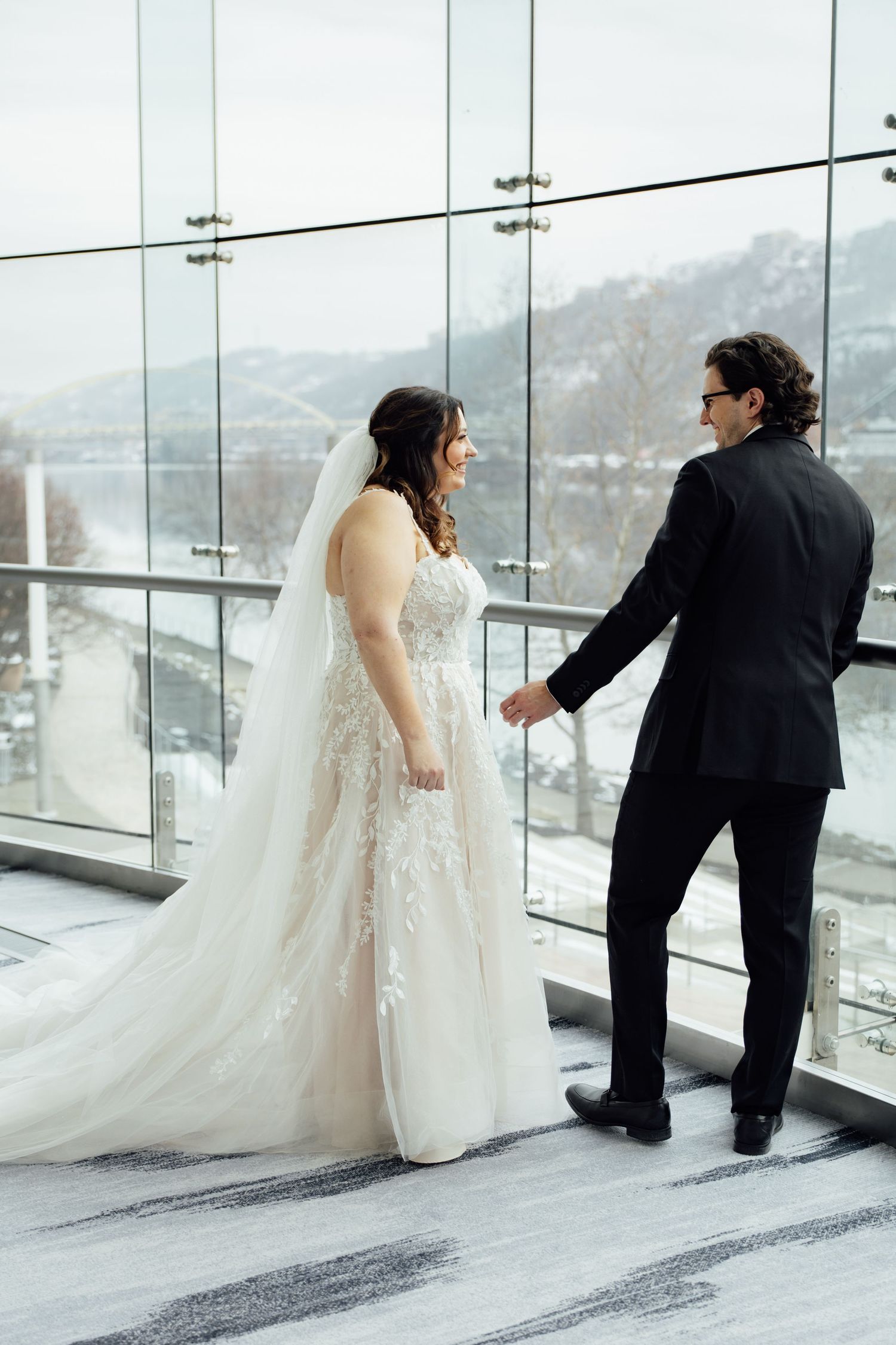 Bride in lace gown and veil holds groom's hand in black suit by glass windows with snowy mountain view.