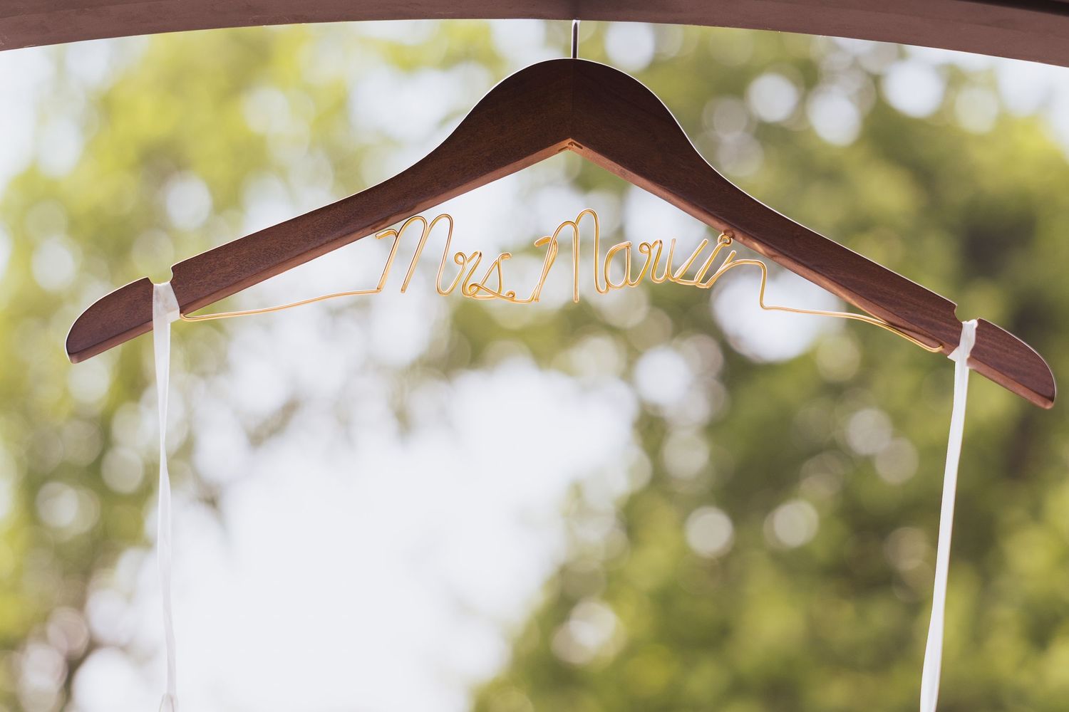 Close-up view of a personalized wooden wedding hanger against a blurred green outdoor background.