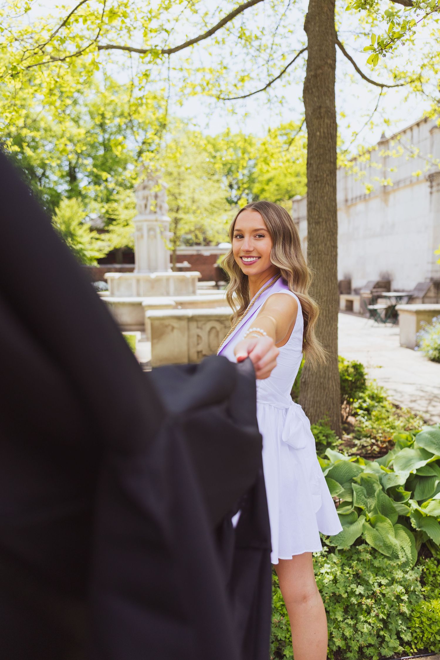 A person in a white summer dress stands by trees and greenery on a sunny day outdoors.
