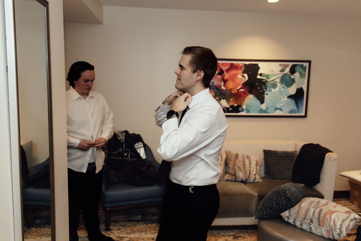 Two men in white dress shirts preparing in a modern hotel room with colorful wall art in the background.