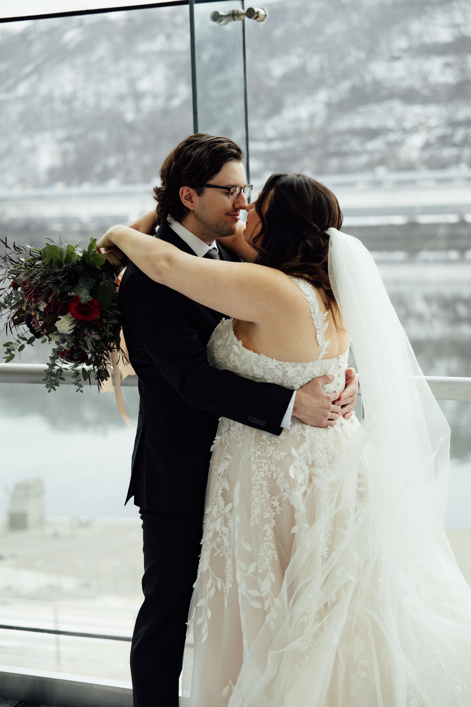 Bride and groom embrace and kiss in front of a snowy mountain view, bride wearing lace gown with veil holding red bouquet.