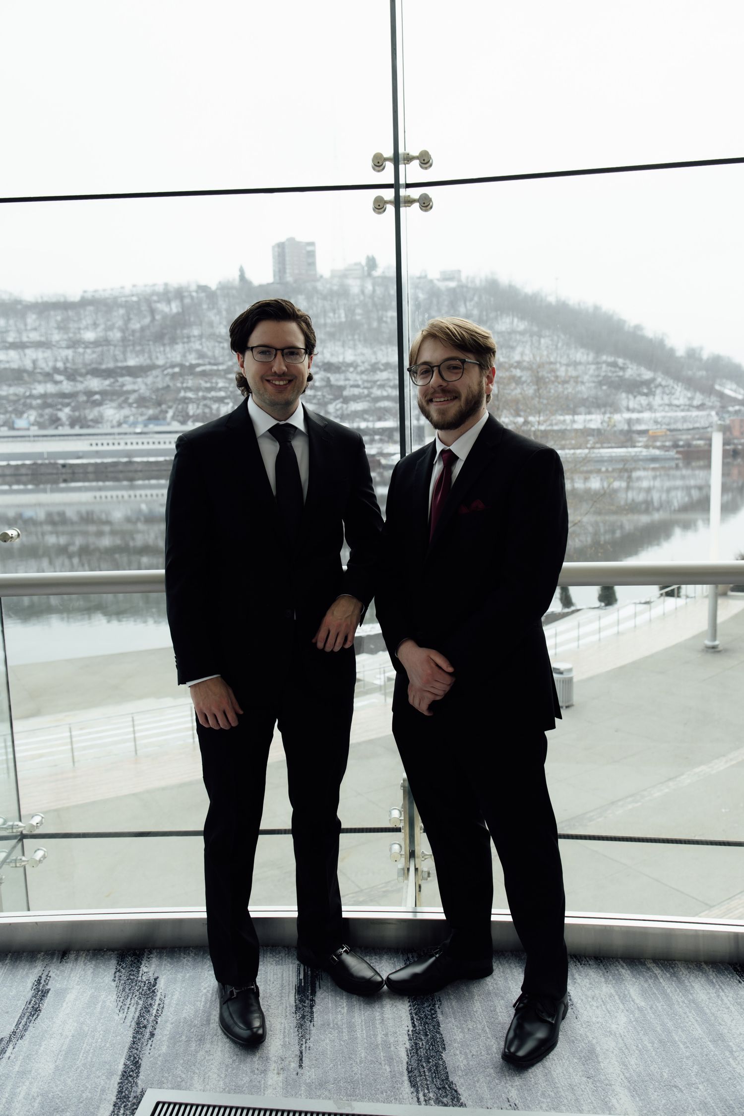Two men in black suits and glasses smiling indoors with a snowy riverside cityscape visible through glass windows.