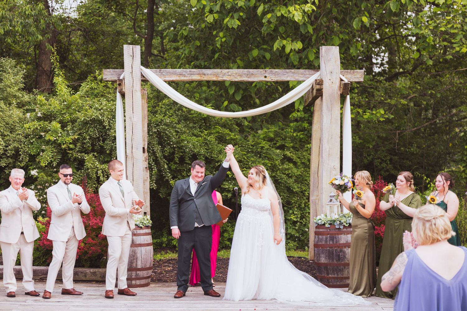 Outdoor wedding ceremony under a white draped arch with wedding party and guests in attendance.
