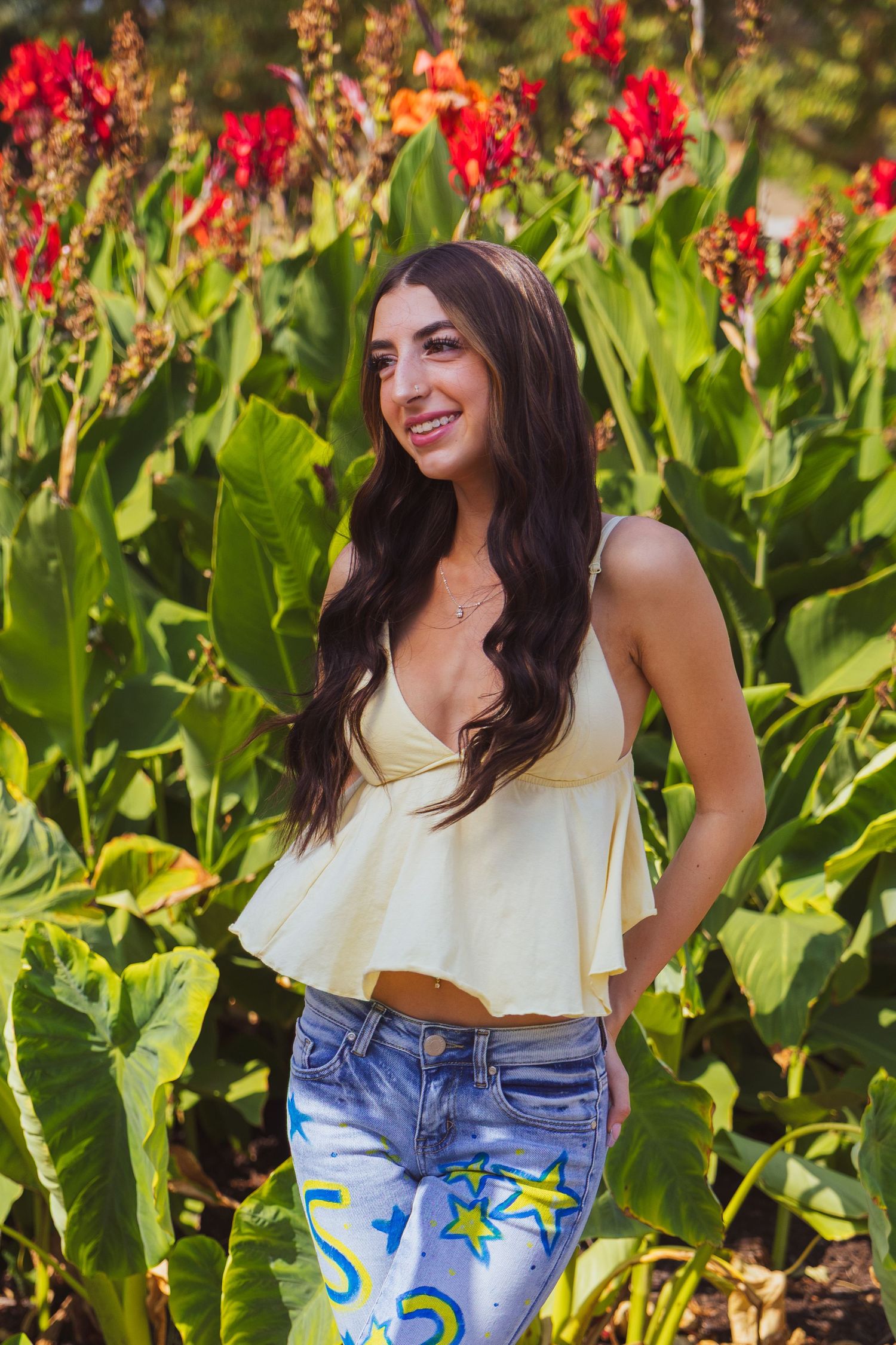 Young woman in yellow crop top and star-print jeans smiling in front of lush green tropical plants with red flowers.