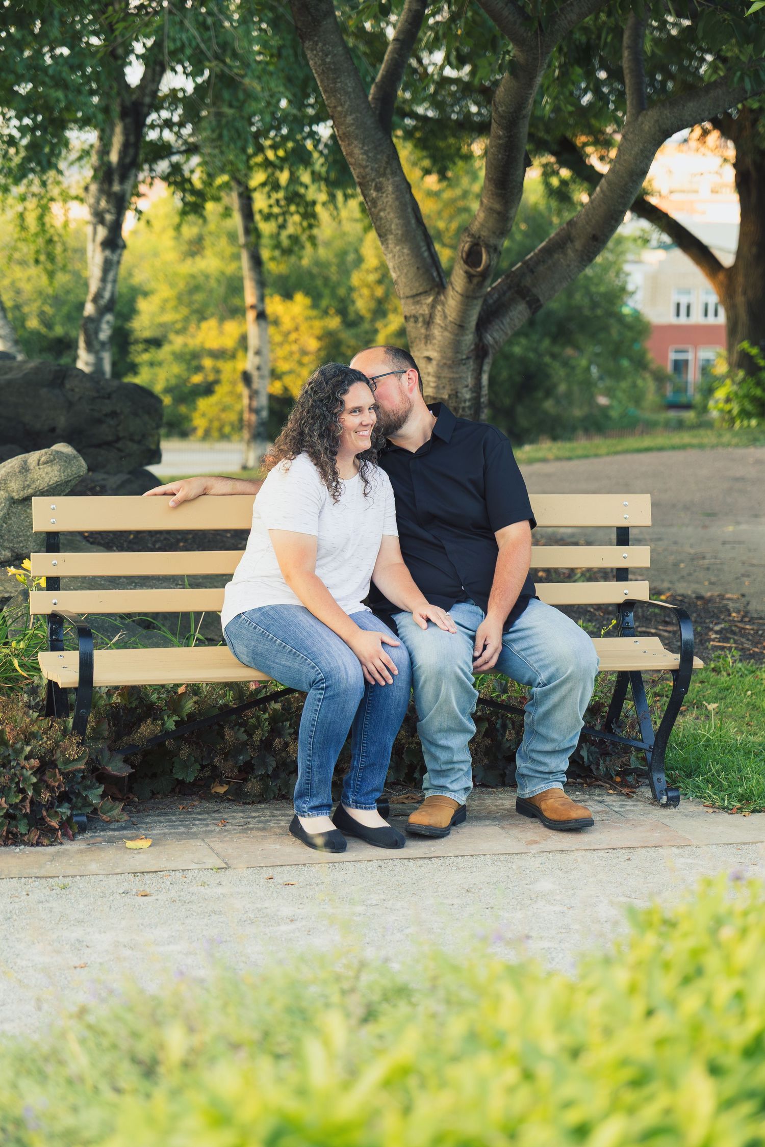 Engaged couple sitting close together on a park bench, smiling and holding hands surrounded by lush green trees.