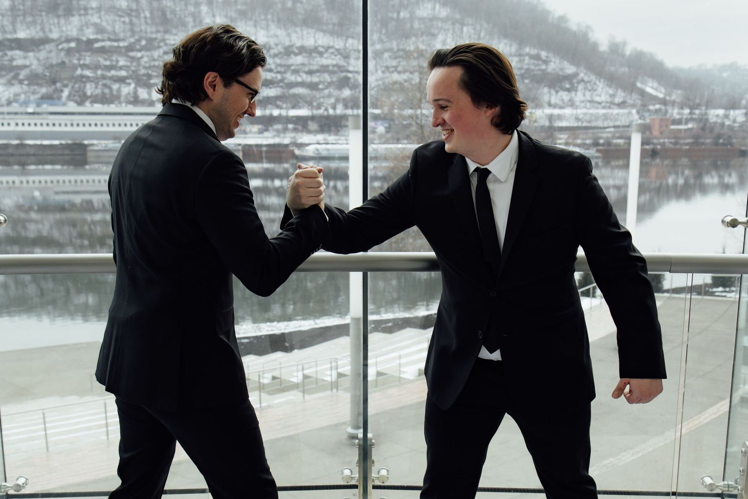 Two men in black suits shaking hands and smiling near a large window with a snowy river view.
