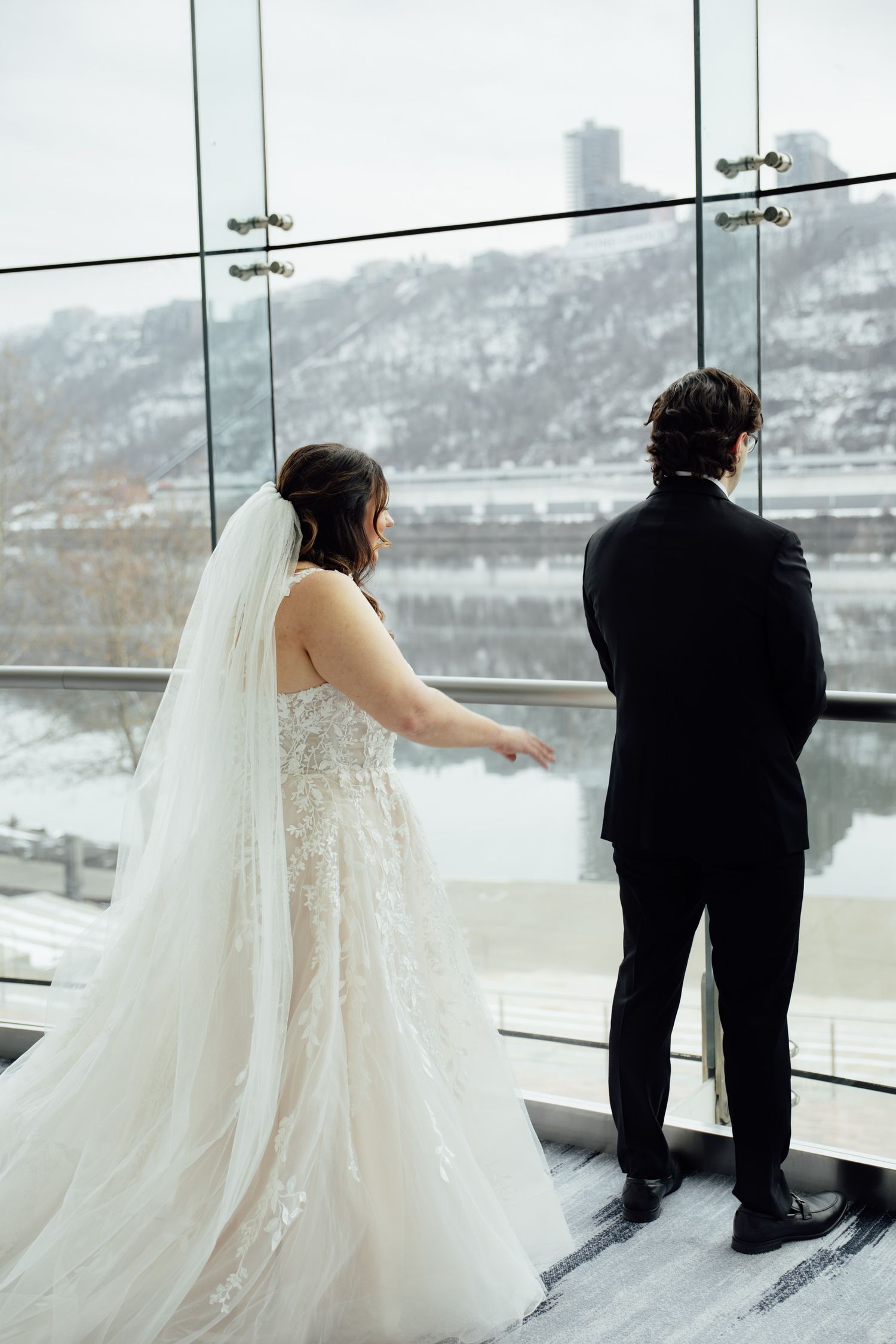 Bride in lace gown with veil and groom in black suit stand before floor-to-ceiling windows with snowy river view.