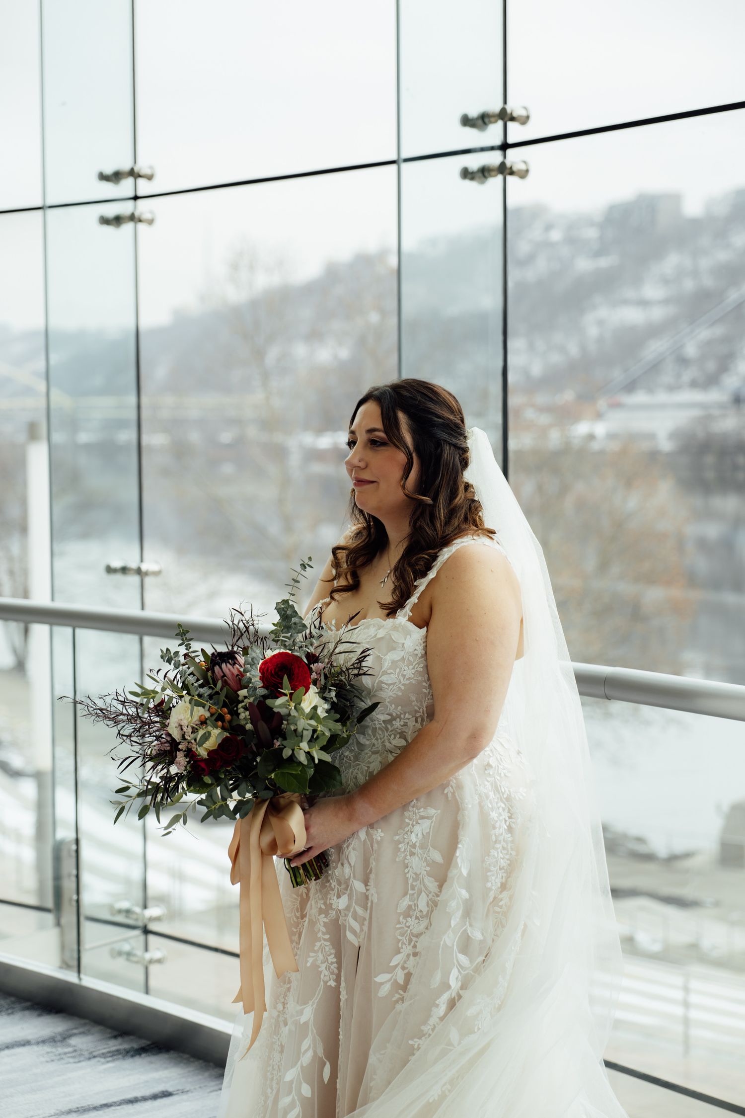 Bride in lace gown holding burgundy bouquet stands before floor-to-ceiling windows with snowy winter landscape backdrop.