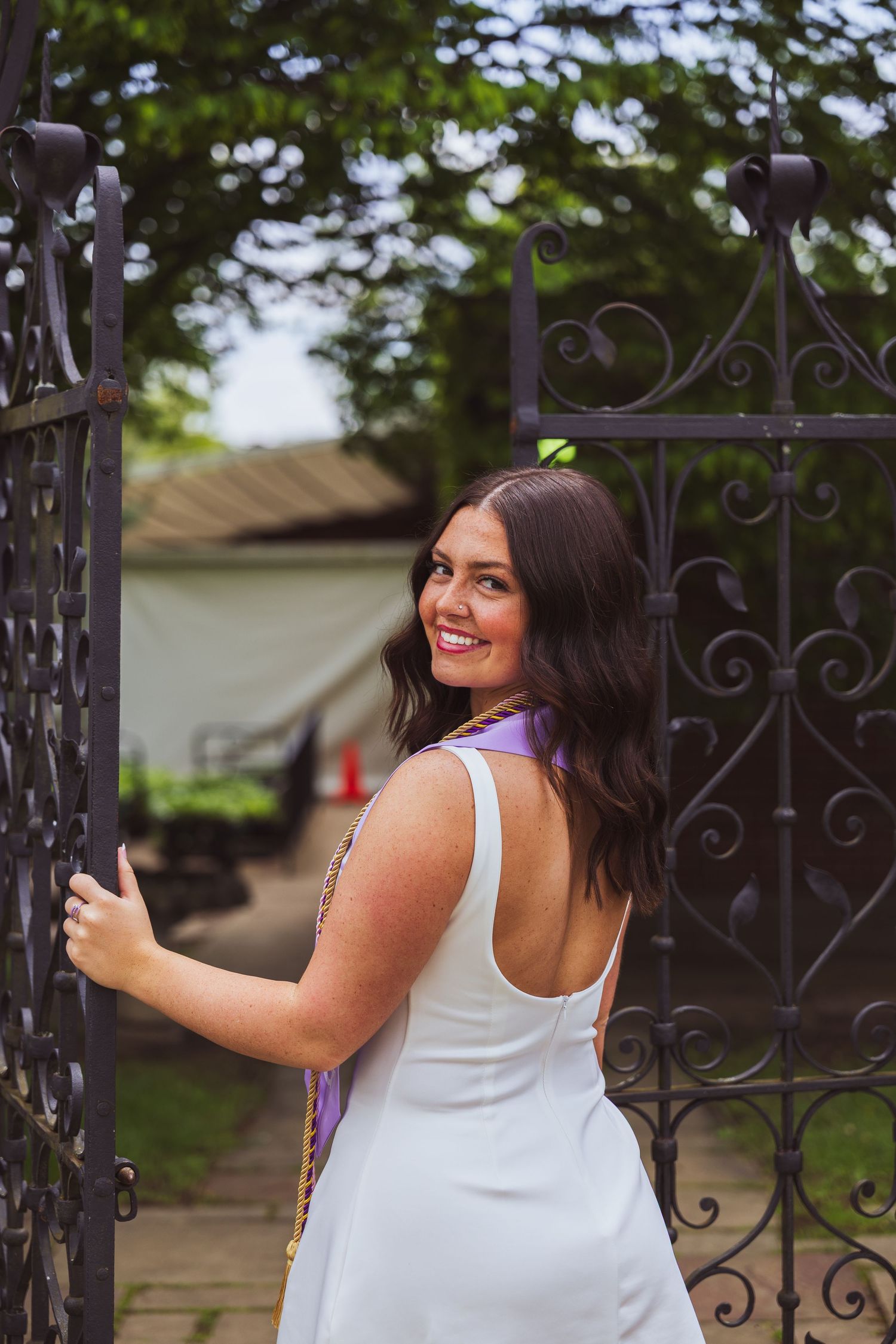A person in a white sundress stands by an ornate black iron garden gate, looking back with a bright smile.
