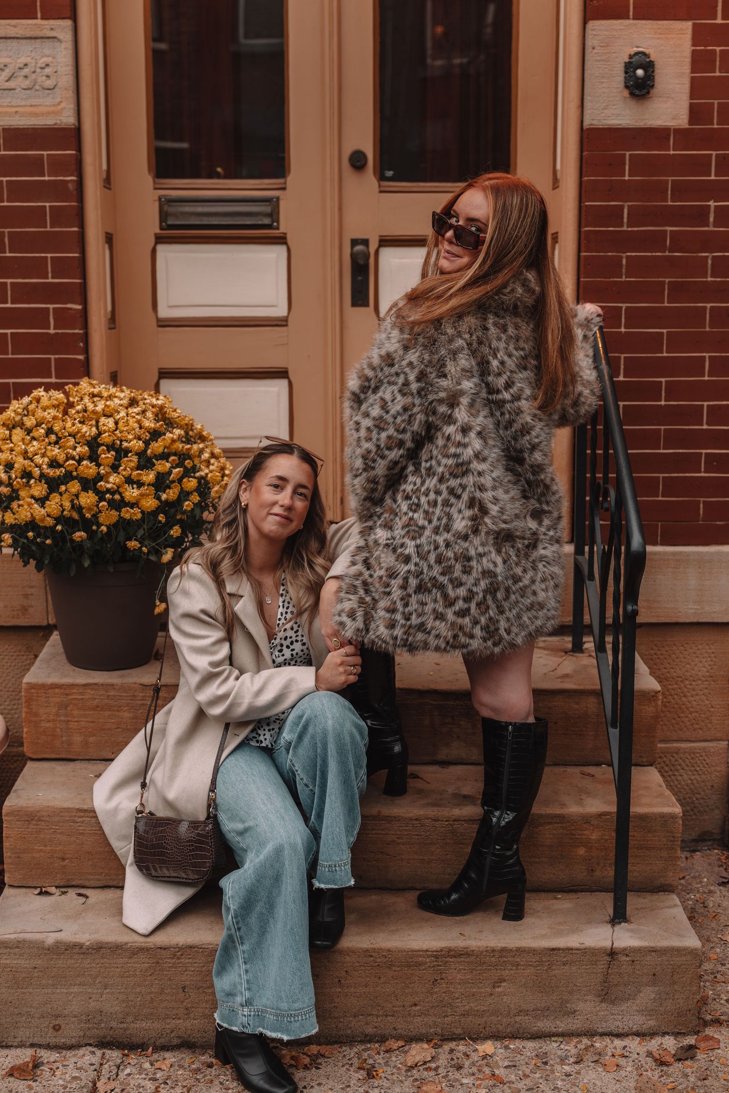 Two women pose on brick building steps in fall fashion, one in cream coat with jeans, other in leopard fur coat with tall boots.
