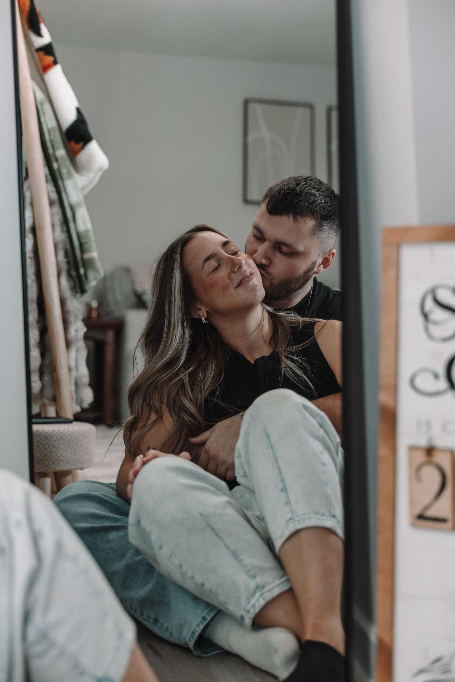 Couple sharing a tender moment, man kissing woman's cheek as she sits between his legs smiling in a cozy bedroom.