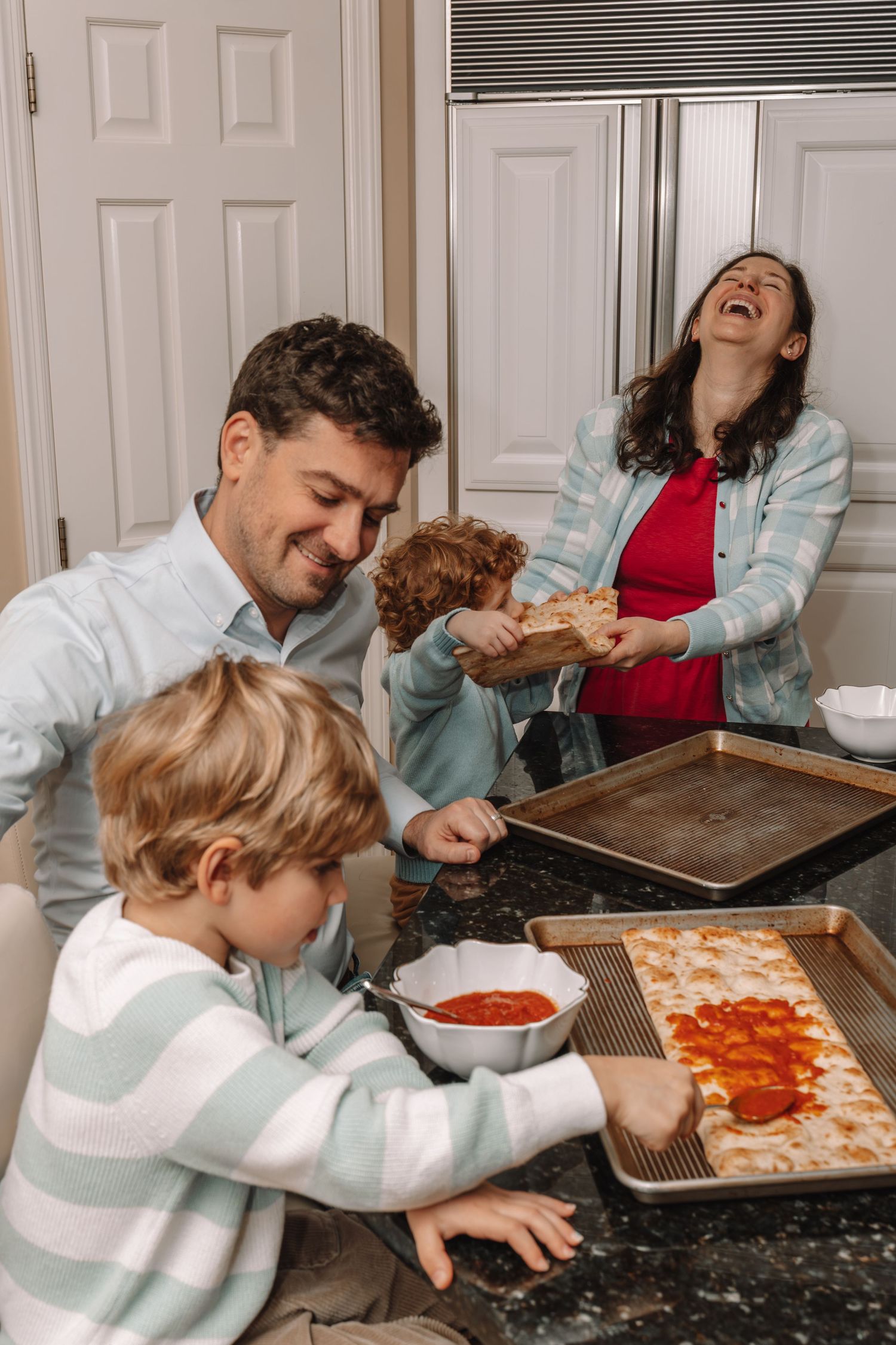Family laughing and making homemade pizza together in a cozy kitchen, with kids helping spread sauce and toppings.