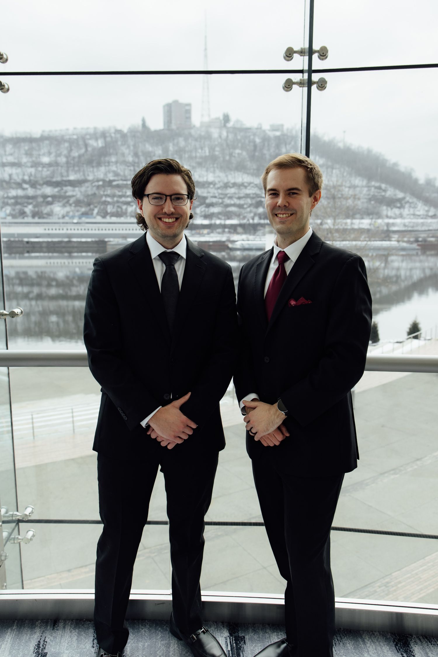 Two men in black suits smiling indoors with a snowy riverside landscape visible through large windows behind them.