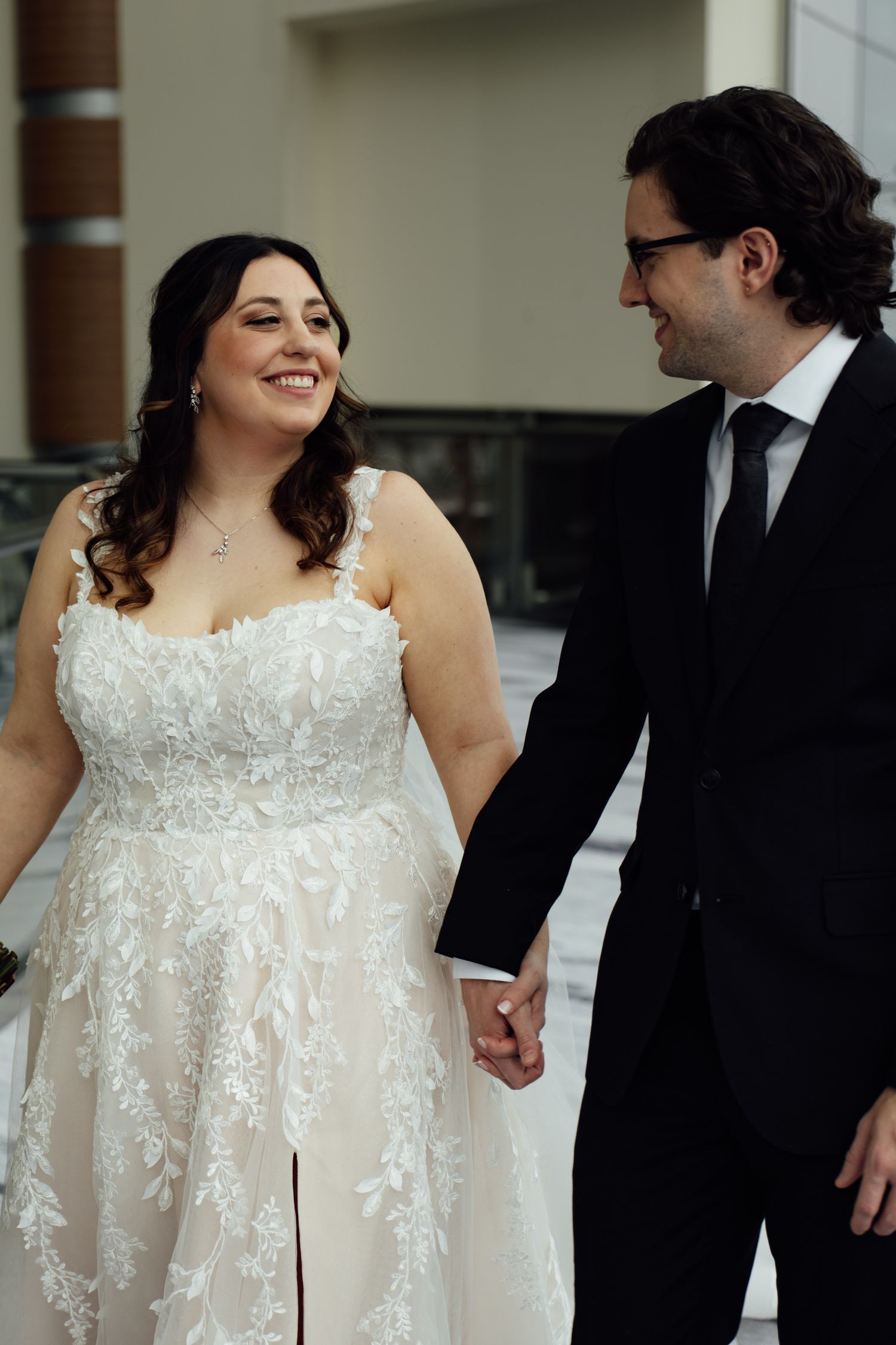 Smiling bride in lace gown holds hands with groom in black suit during outdoor winter wedding.