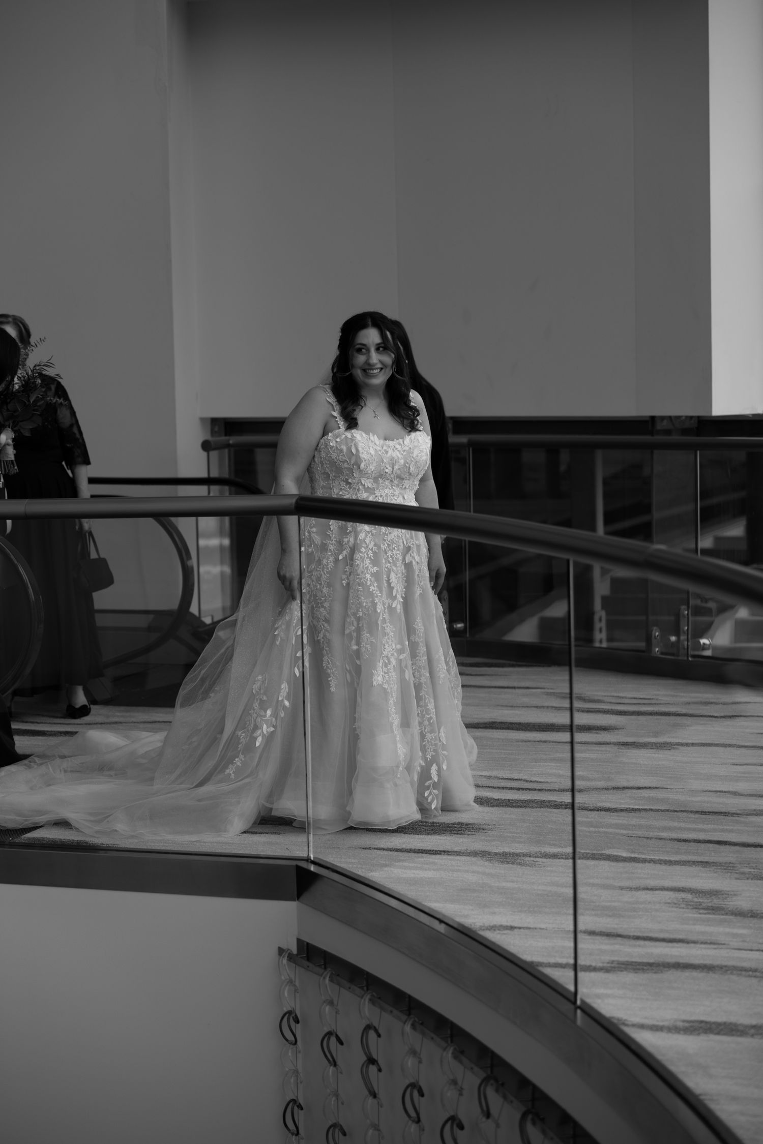 Bride in floral lace ball gown poses on curved staircase balcony in elegant black and white wedding photo.