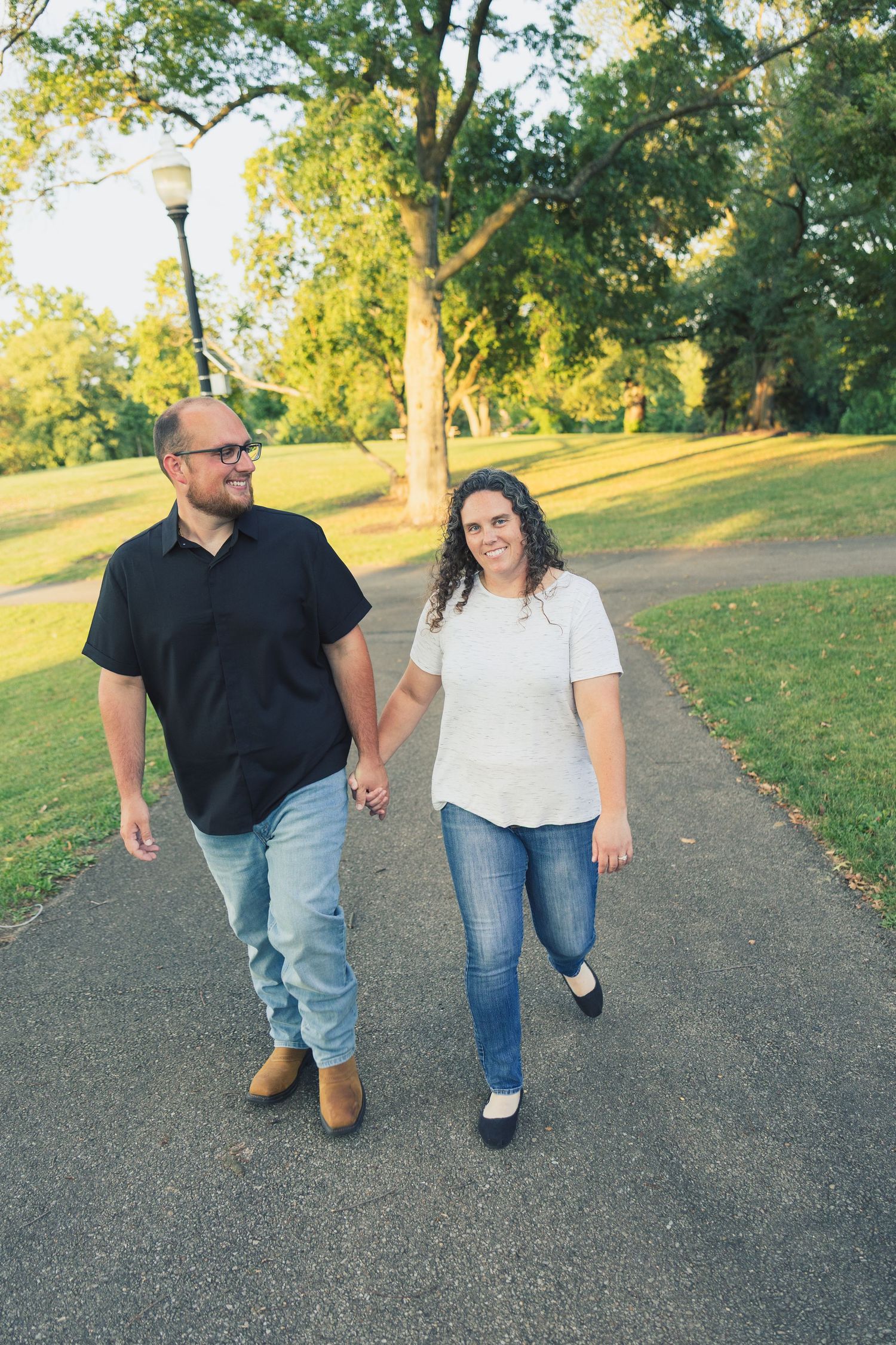 Happy couple holding hands walking on park path, man in black shirt and woman in white top and jeans.