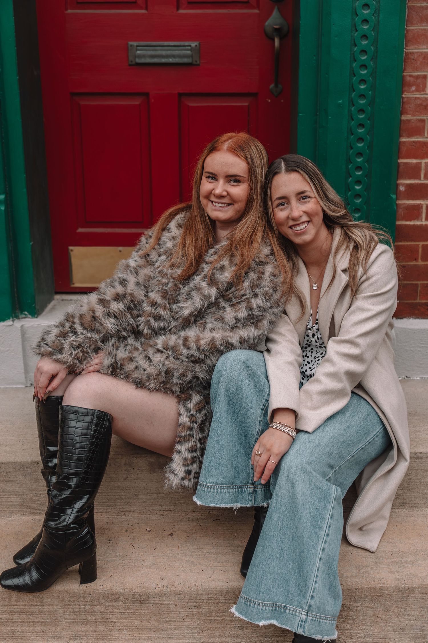 Two women smiling on red-doored steps, one in faux fur coat with black boots, other in beige blazer and wide-leg jeans.