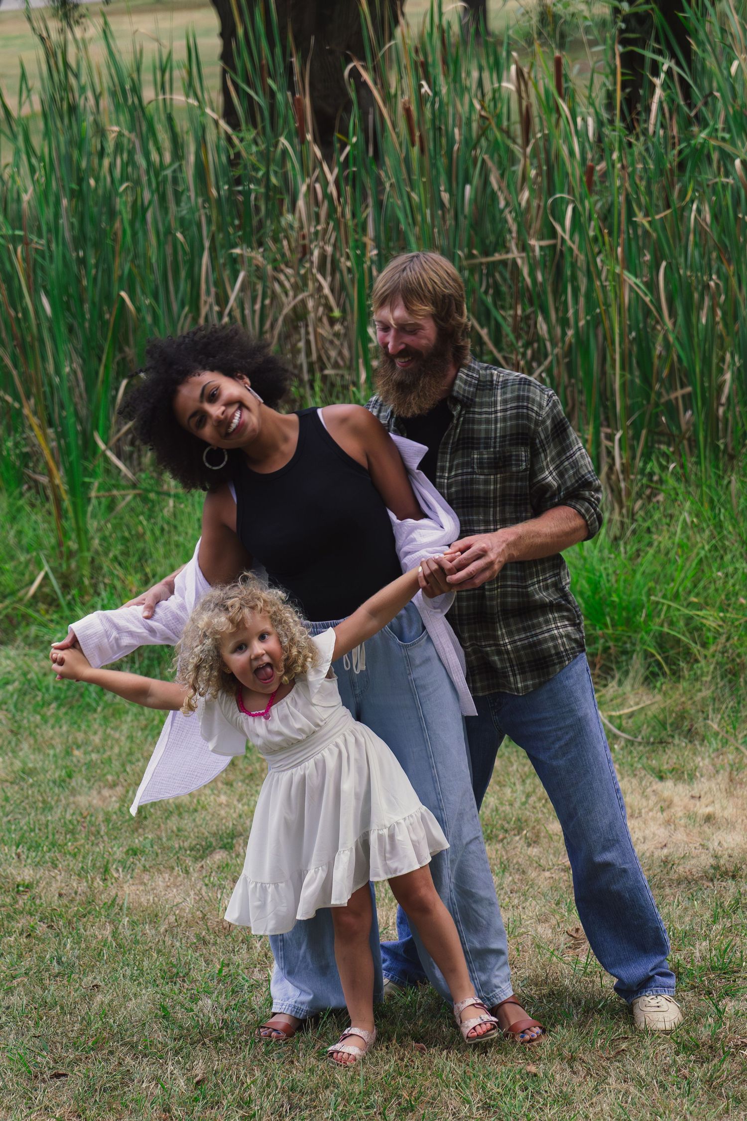 A joyful multiracial family plays outdoors near tall reeds, with a laughing curly-haired girl in a white dress.