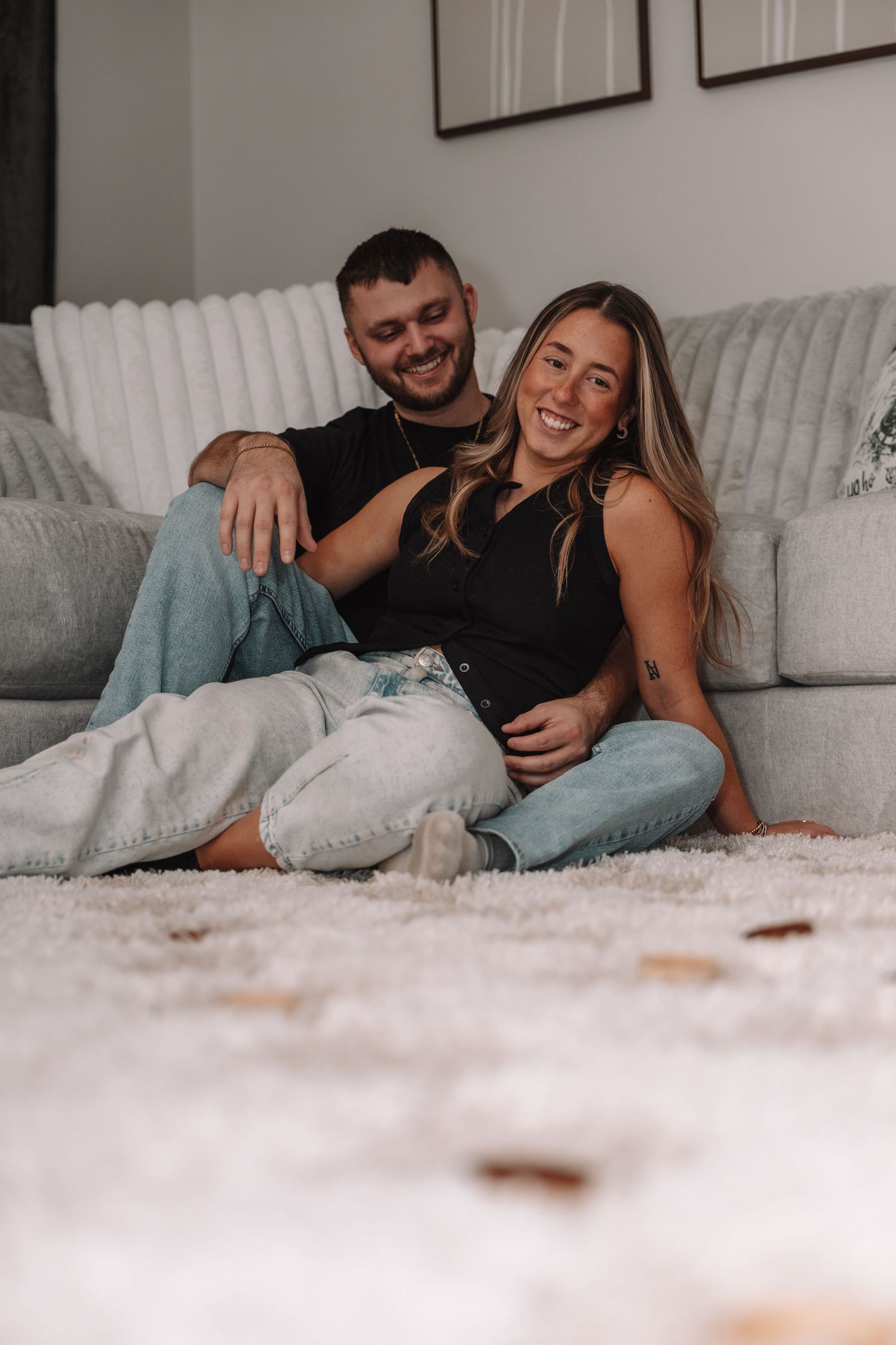 Happy couple sitting on a white fluffy carpet leaning against a gray couch, smiling and embracing indoors.