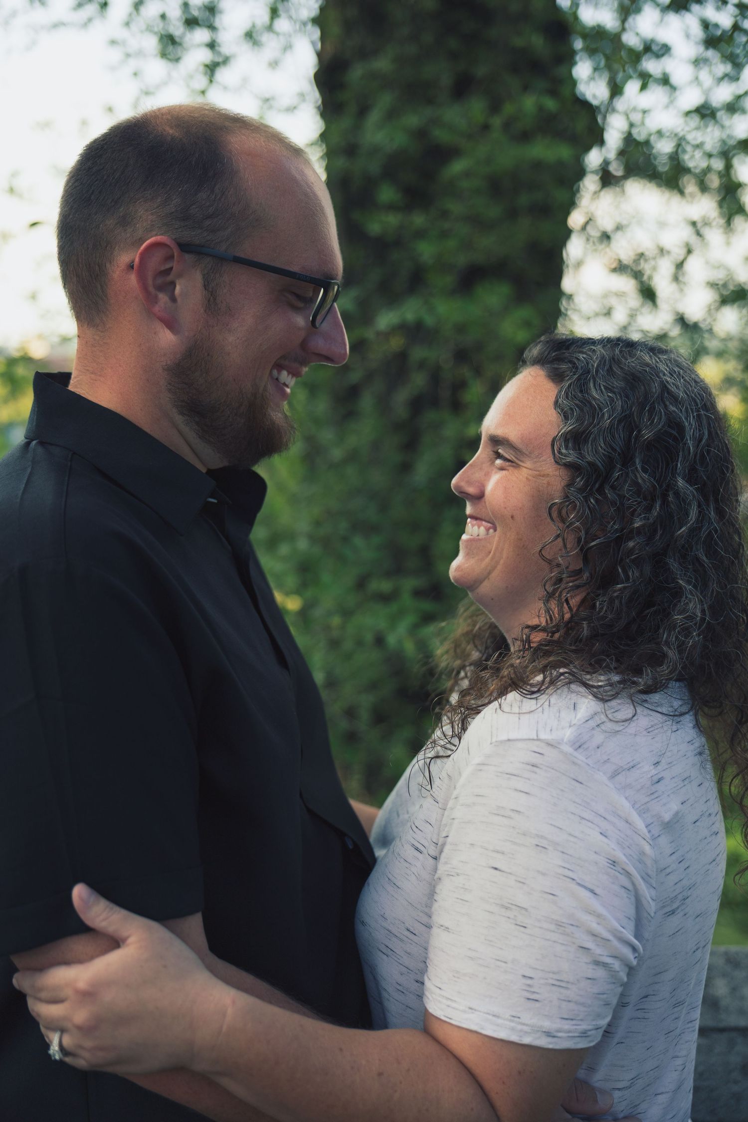 Smiling couple embracing outdoors, man in black shirt with glasses and woman with curly hair in white top.