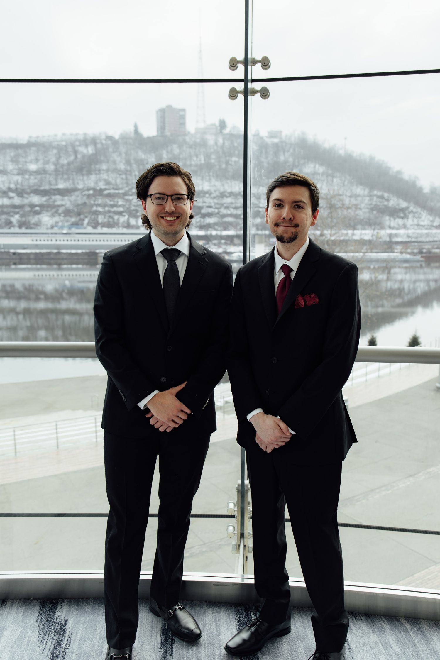 Two young men in black suits smiling indoors with a snowy cityscape visible through large windows behind them.