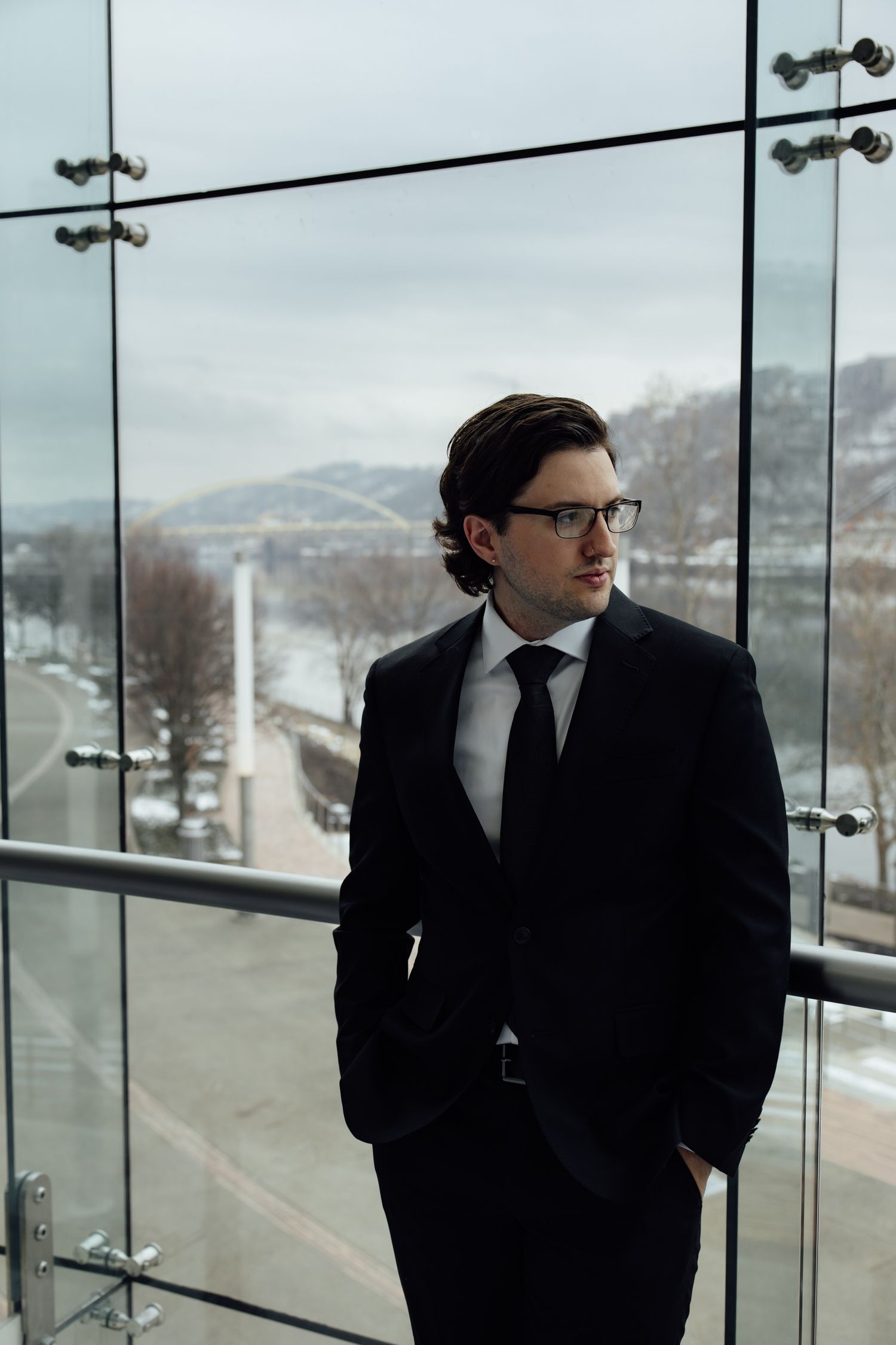 Stylish businessman in dark suit with hands in pockets on glass office balcony.