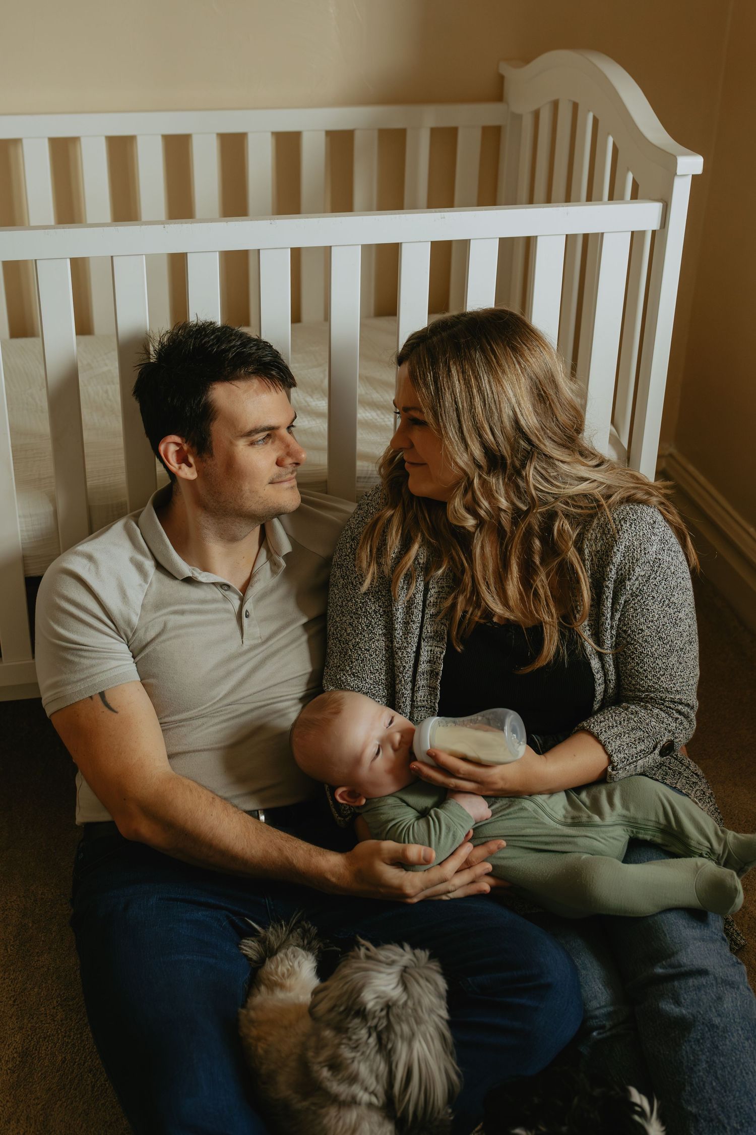 A young couple sits on the floor by a white crib, feeding their newborn baby a bottle with a dog nearby.
