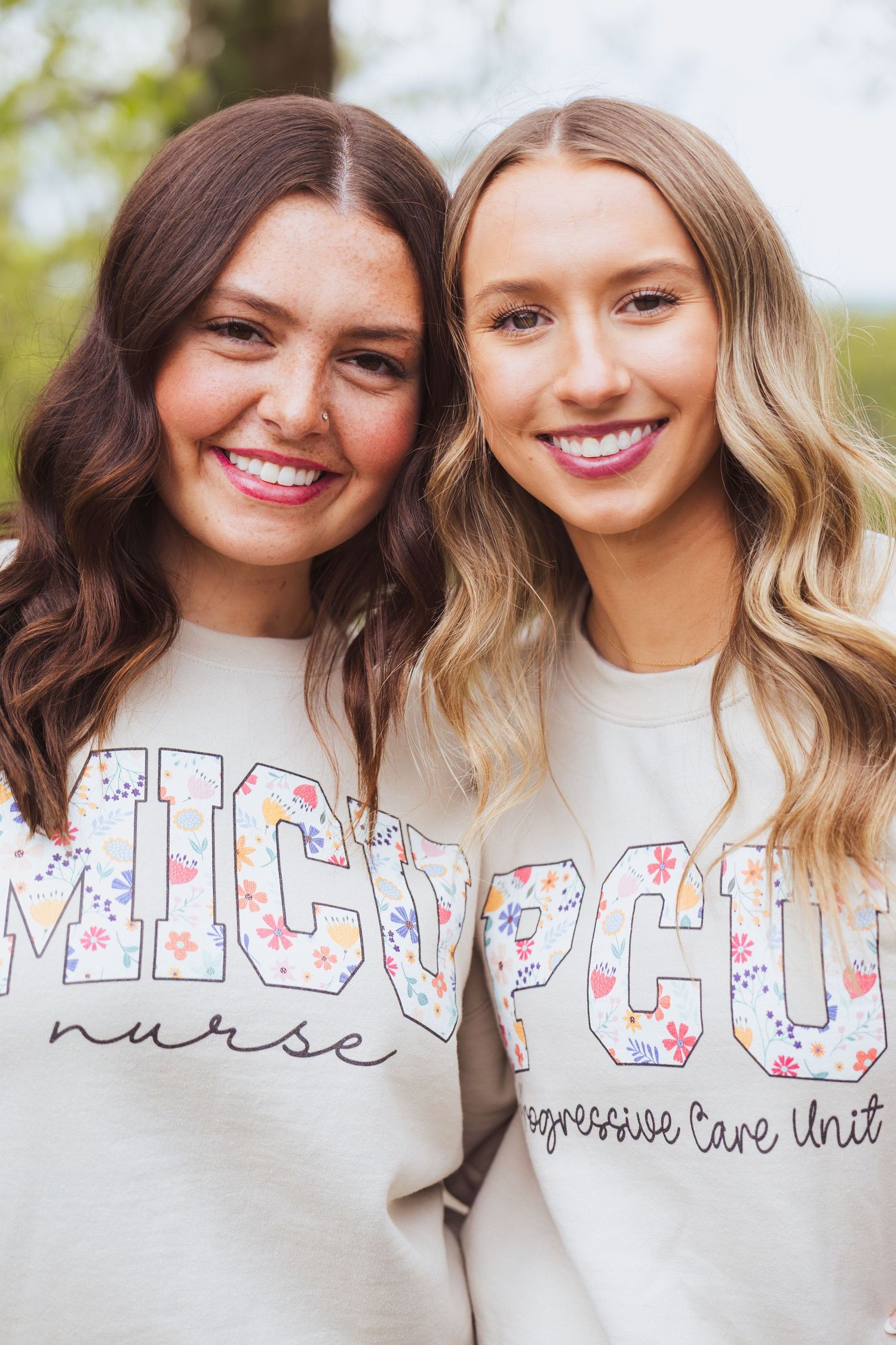 Two nurses wearing matching floral ICU sweatshirts smile together outdoors.