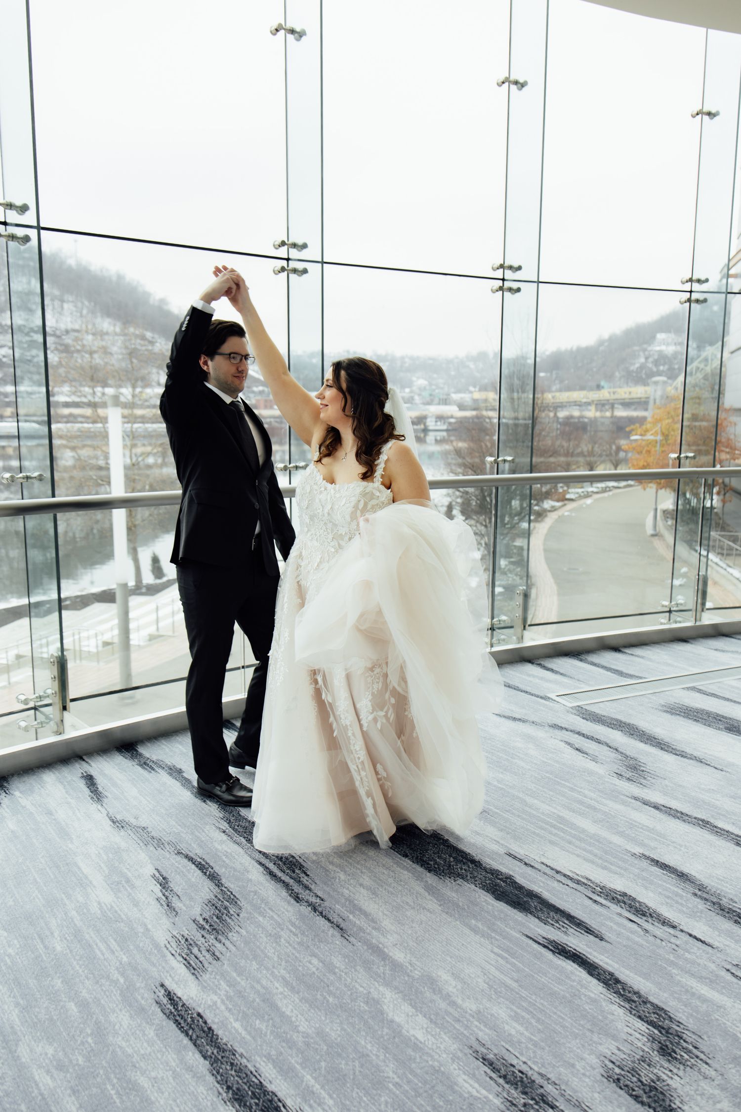 A bride and groom share a romantic dance in a modern ballroom with floor-to-ceiling windows overlooking a snowy winter landscape.