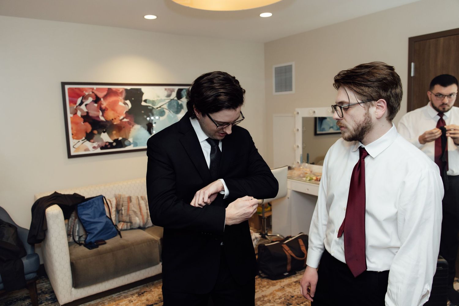 Two men in formal attire preparing for wedding, one in black suit checking watch in elegant hotel suite.