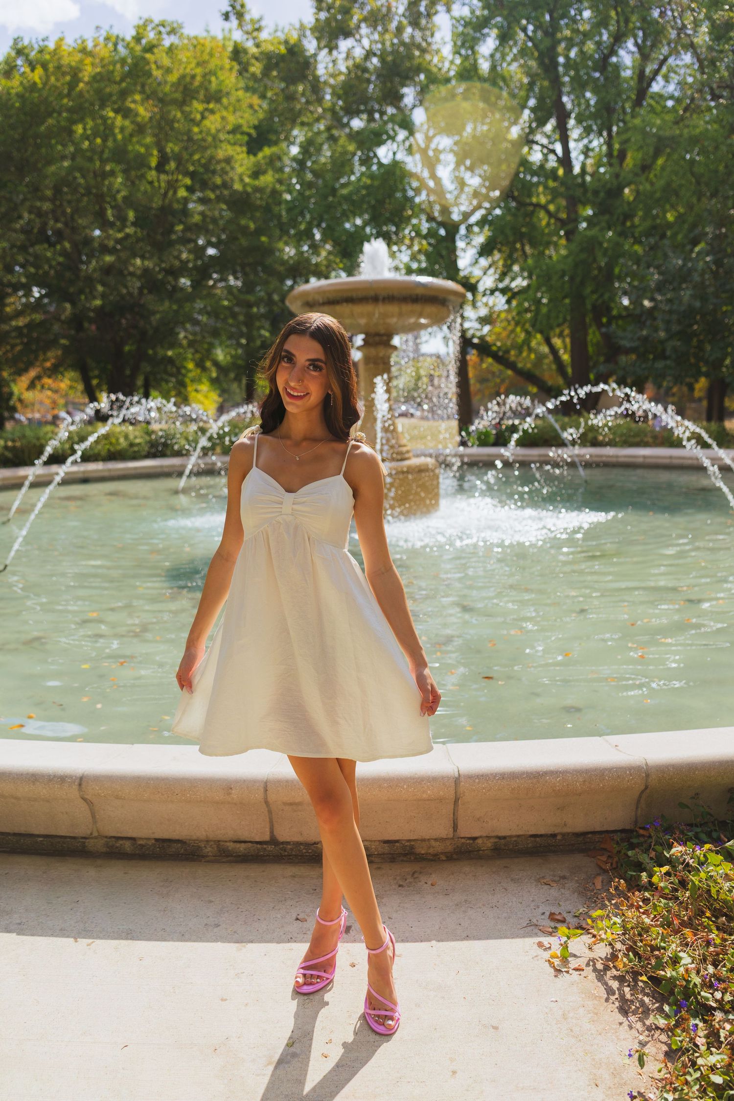 Young woman in white mini dress and pink heels posing by a decorative fountain in a sunny park.