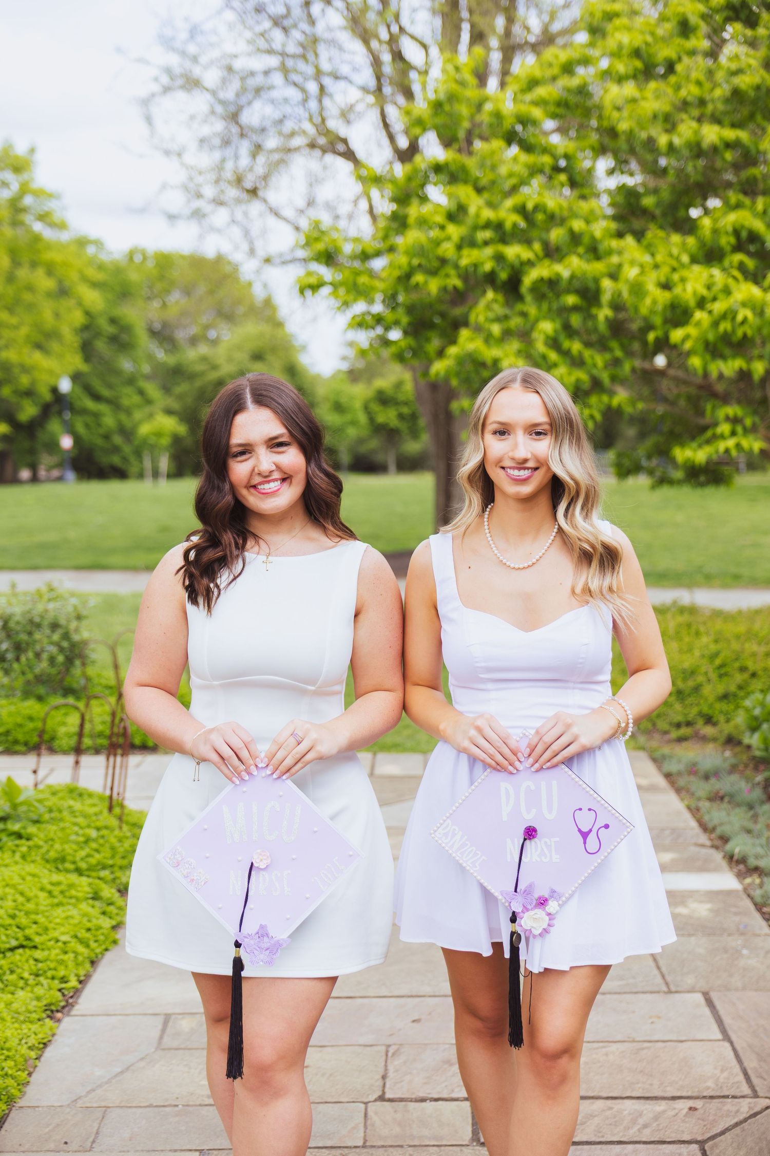 Two women in white dresses pose with golf clubs on a garden path surrounded by green trees.