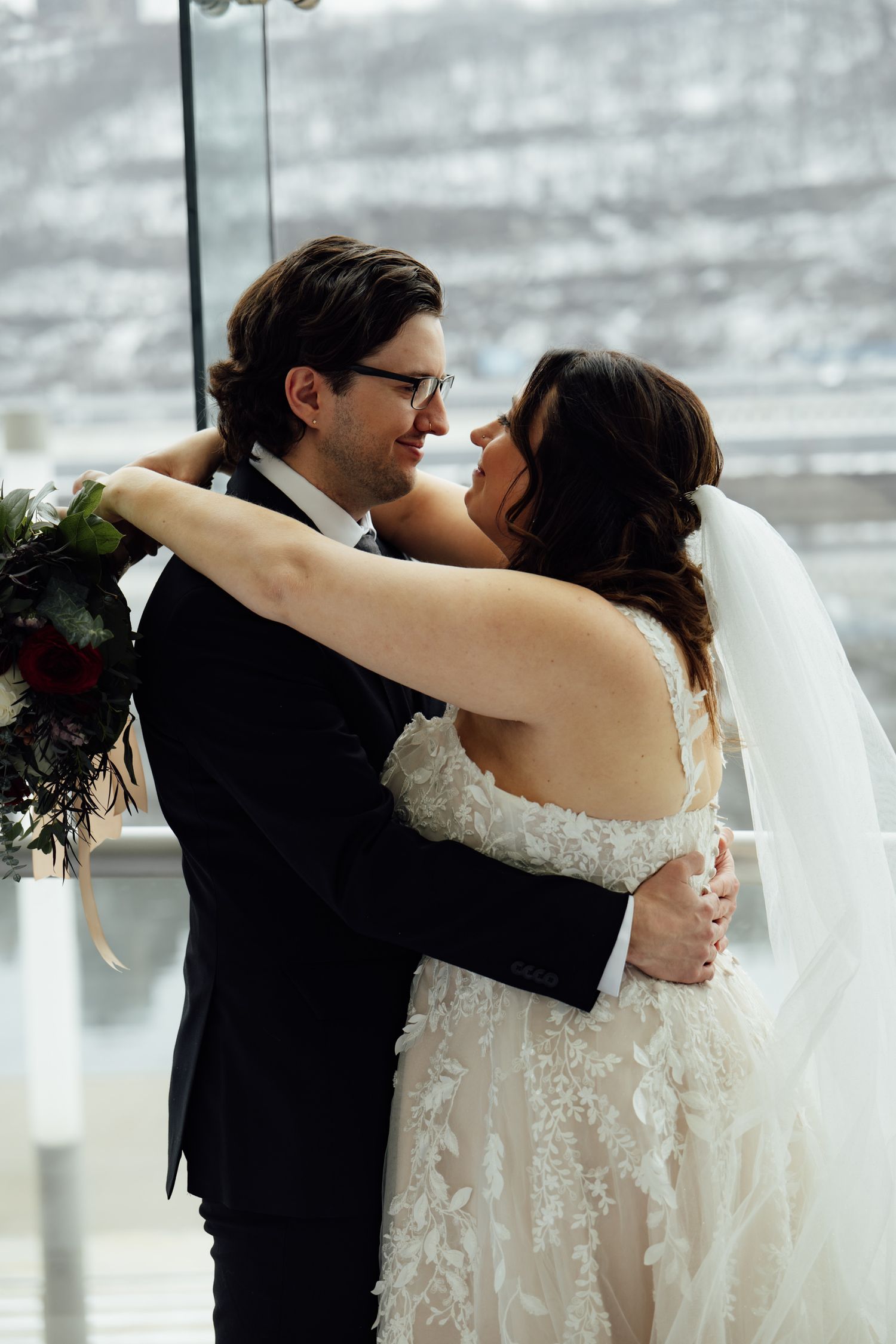 Bride and groom embrace tenderly in wedding attire, she in lace gown with veil, he in black tuxedo, near large windows.