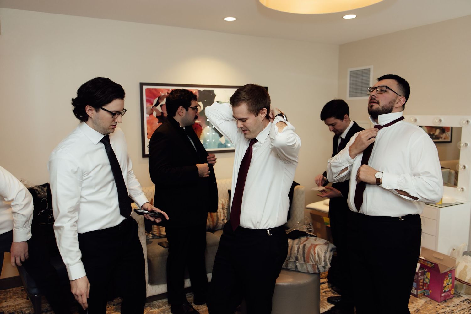 Groomsmen in white shirts and dark ties help each other get dressed and adjust ties before a wedding ceremony.