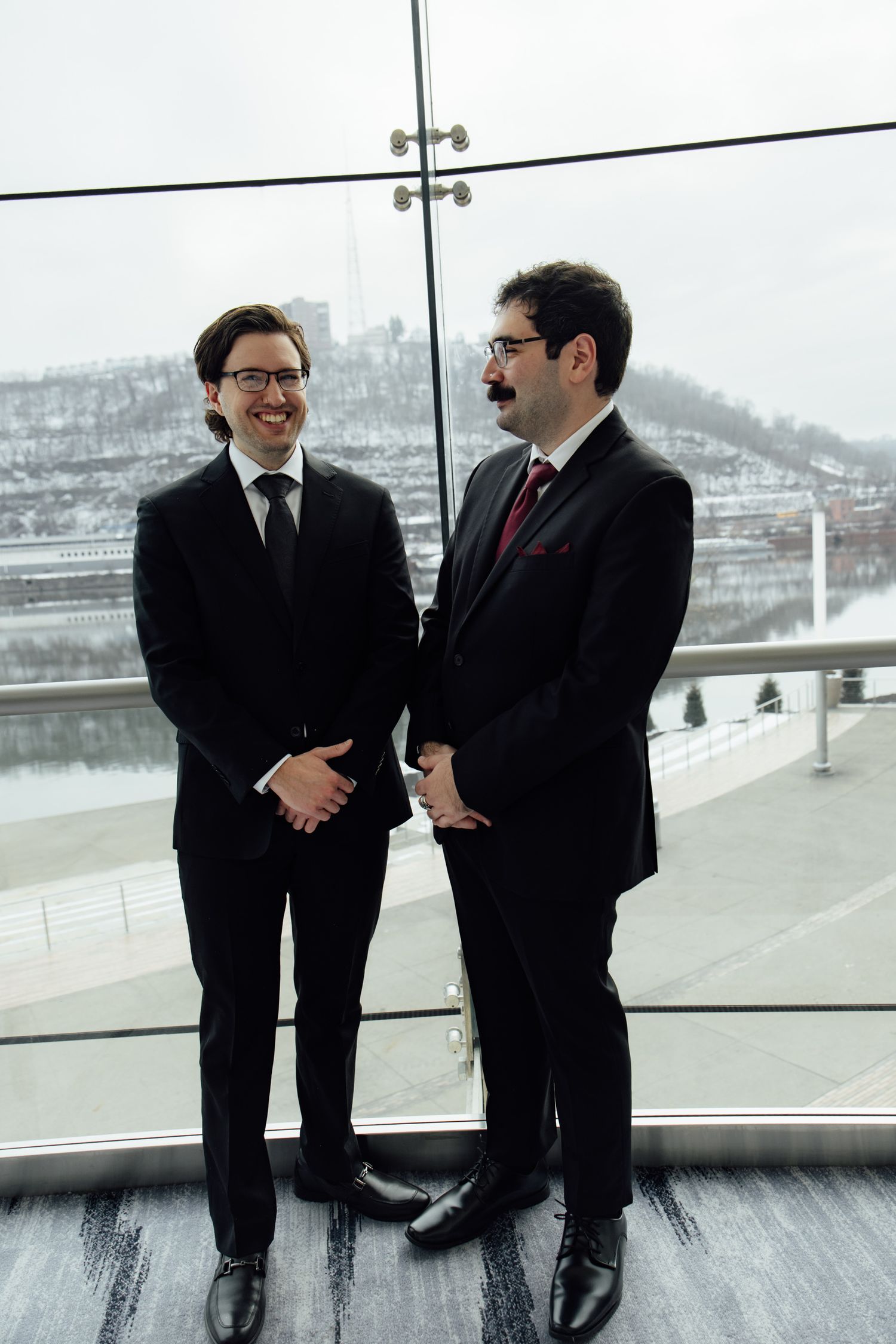 Two professional men in black suits stand smiling by large windows with a snowy cityscape backdrop.