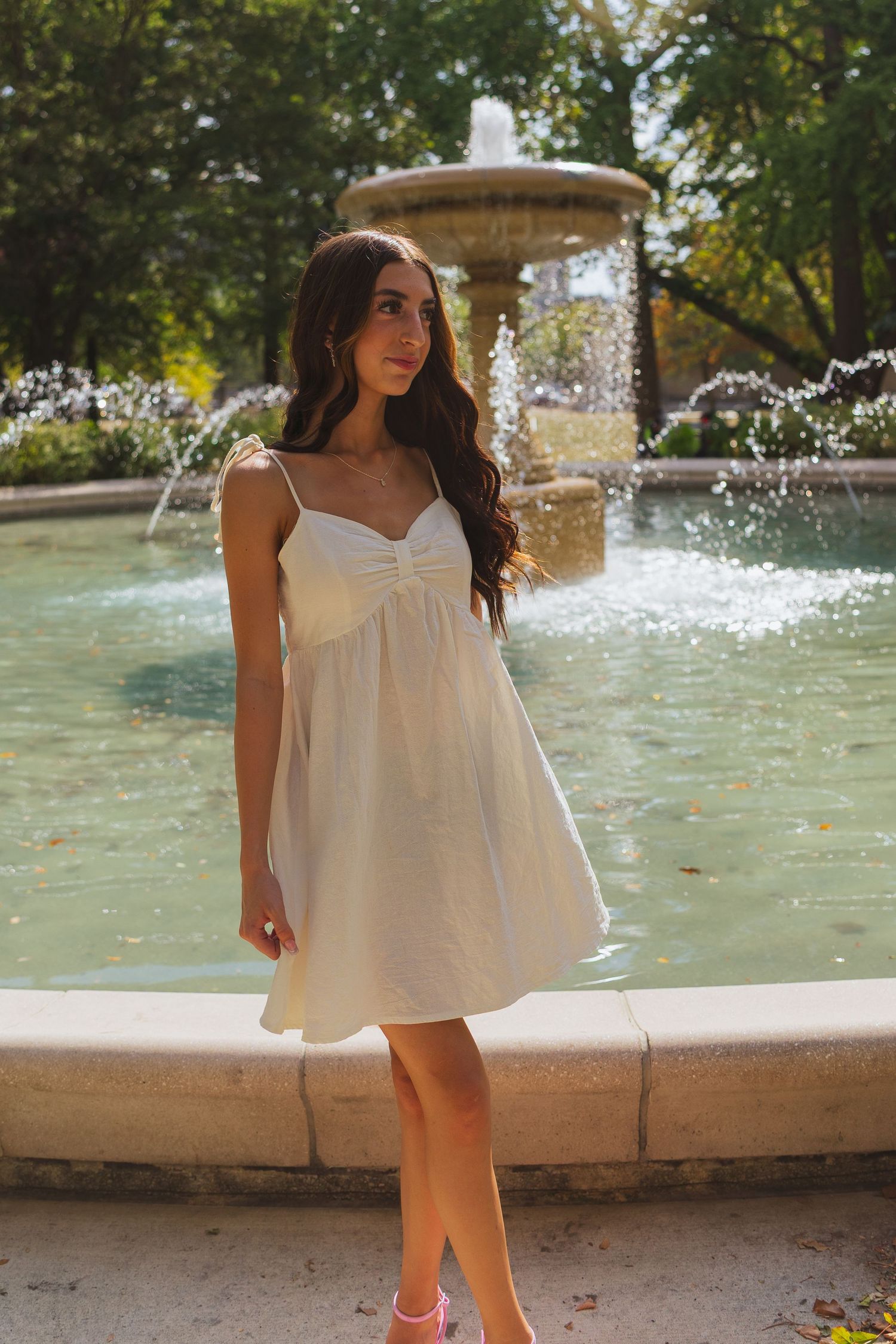 Young woman in white sundress posing by a stone fountain in a lush green park on a sunny day.