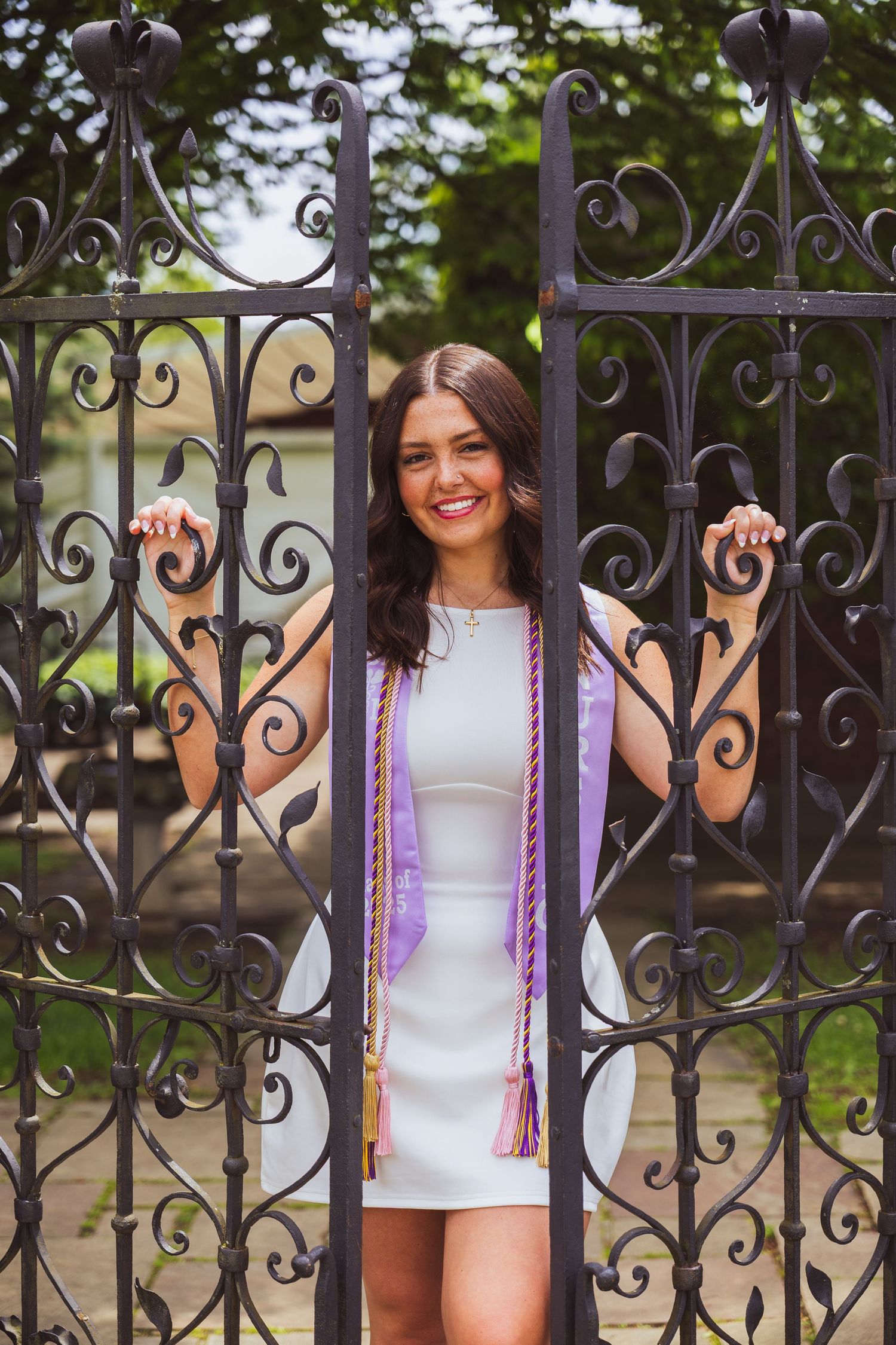 Person standing between ornate wrought iron gates wearing a white dress and purple graduation stole.