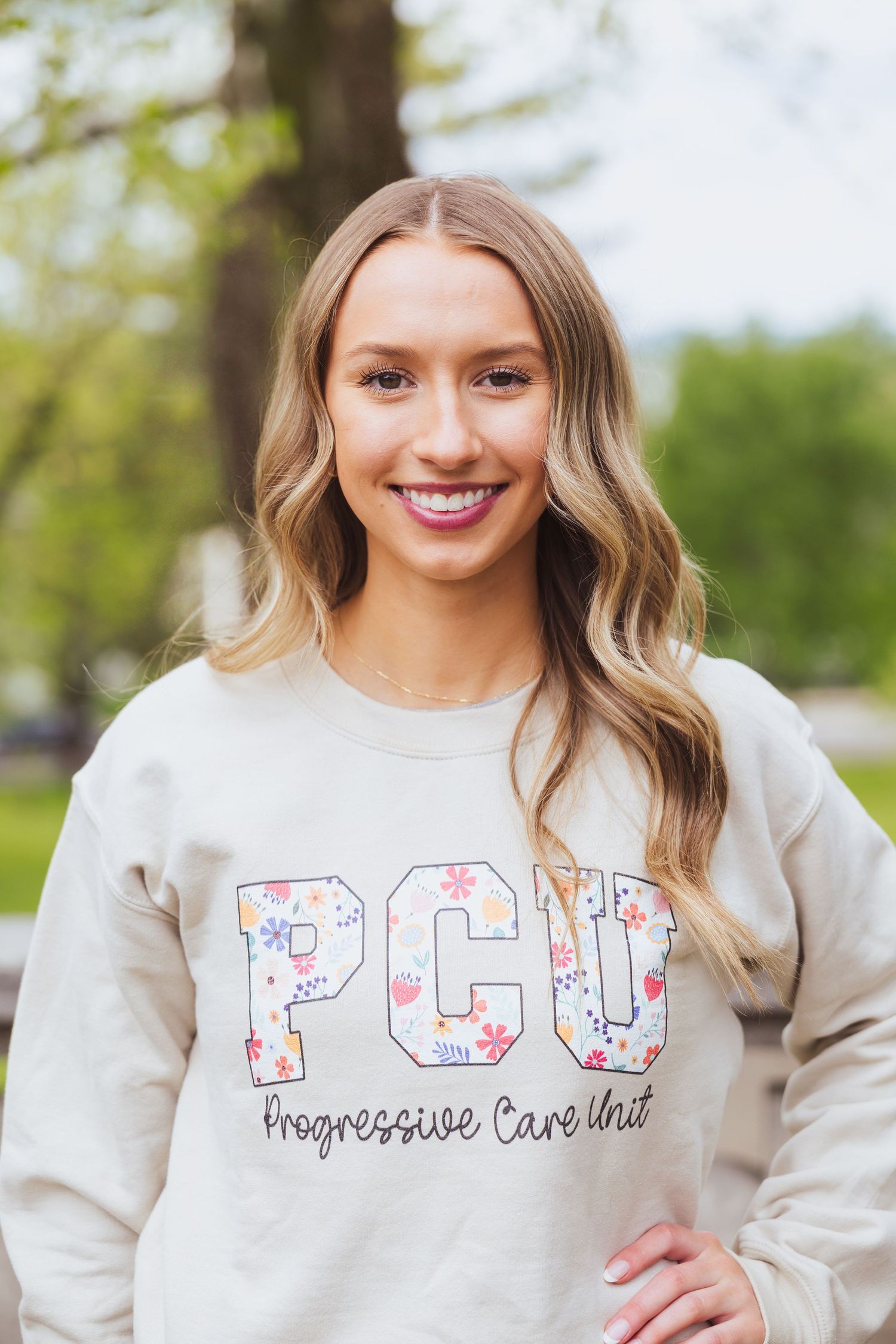 A person wearing a floral-patterned PCU lettering cream sweatshirt poses outside in a natural setting.