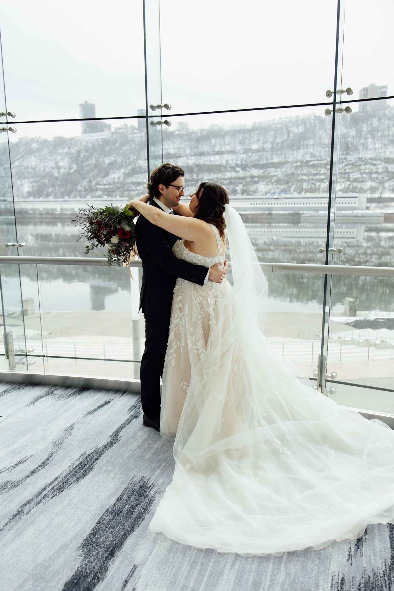 Bride and groom embrace near floor-to-ceiling windows with snowy river views, she holds a red bouquet in a layered white gown.