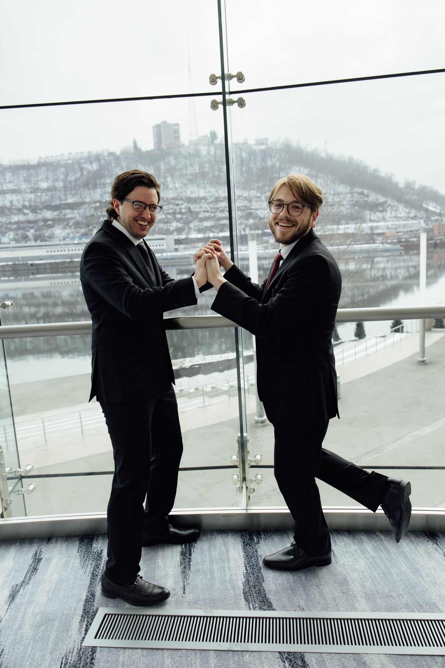 Two smiling businessmen in black suits holding hands on a glass balcony with a snowy cityscape backdrop.