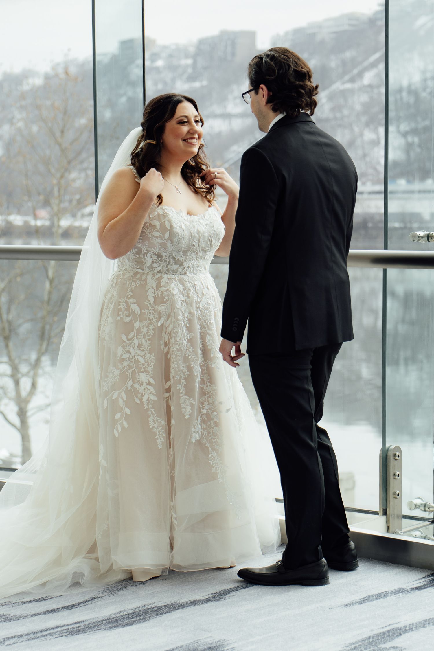 Bride in floral lace ball gown smiles at groom in black suit, standing by floor-to-ceiling windows with snowy backdrop.