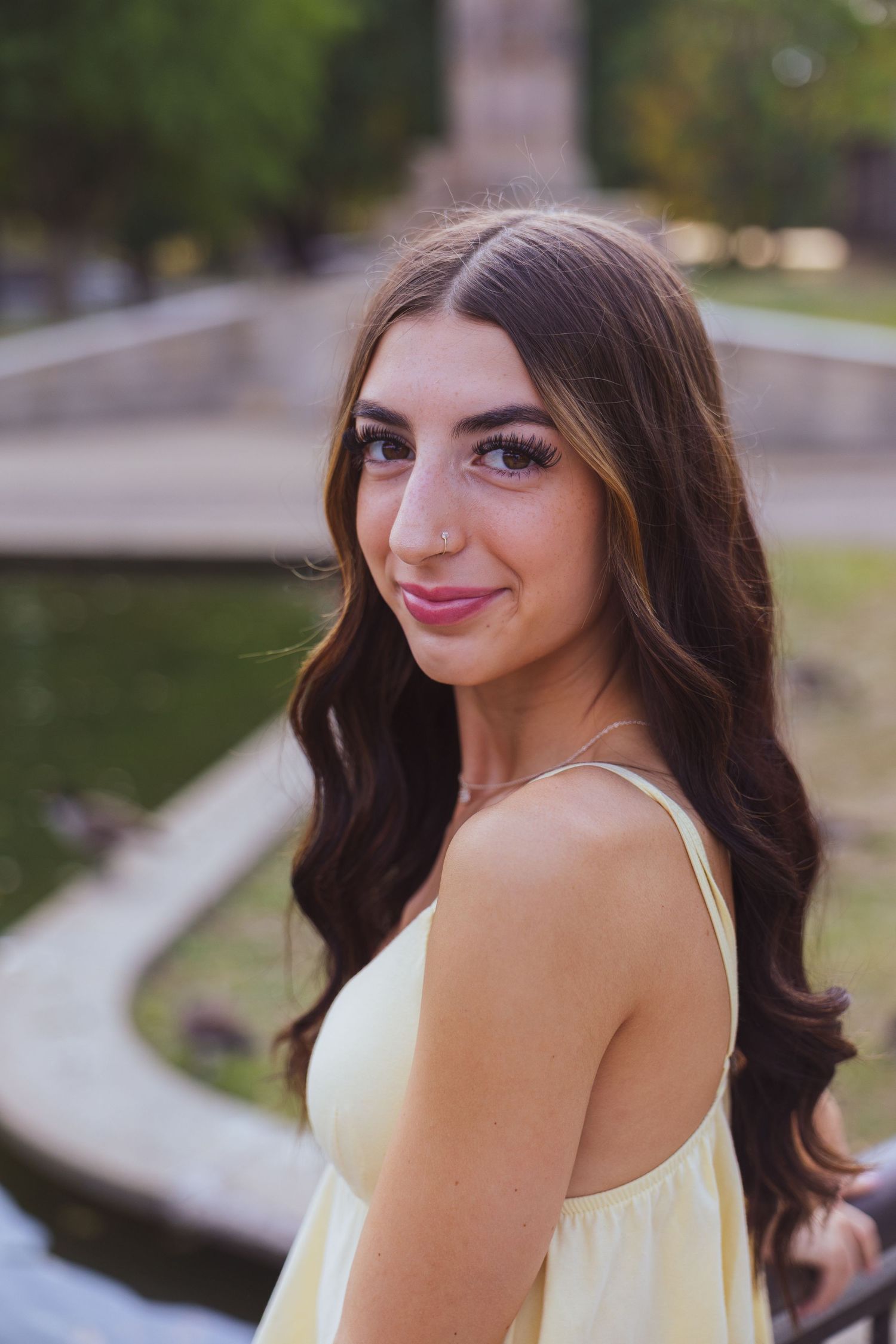 Young woman with long dark wavy hair wearing a yellow sundress, smiling outdoors near a scenic waterway.