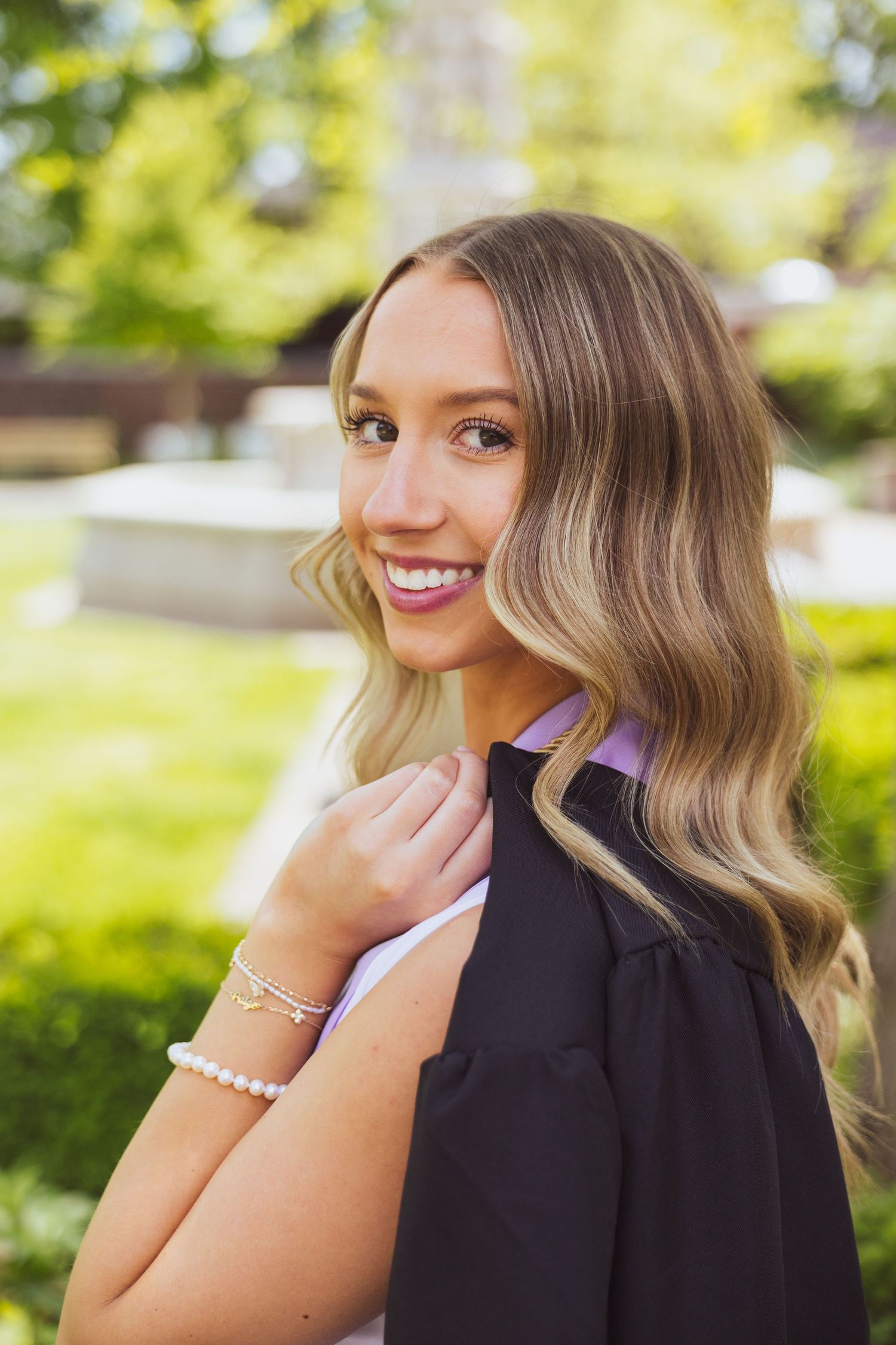 Portrait of a smiling blonde woman with wavy hair in black dress outdoors with sunlit foliage in background.