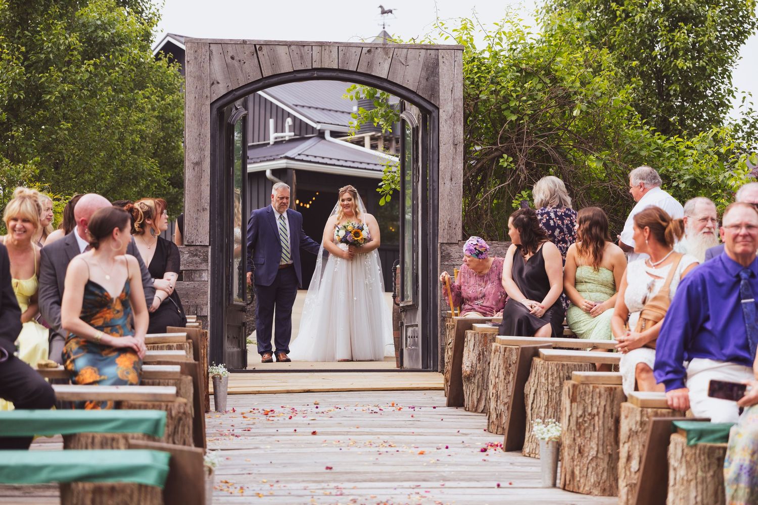 Wedding ceremony guests seated on wooden logs as couple stands under rustic wooden archway.