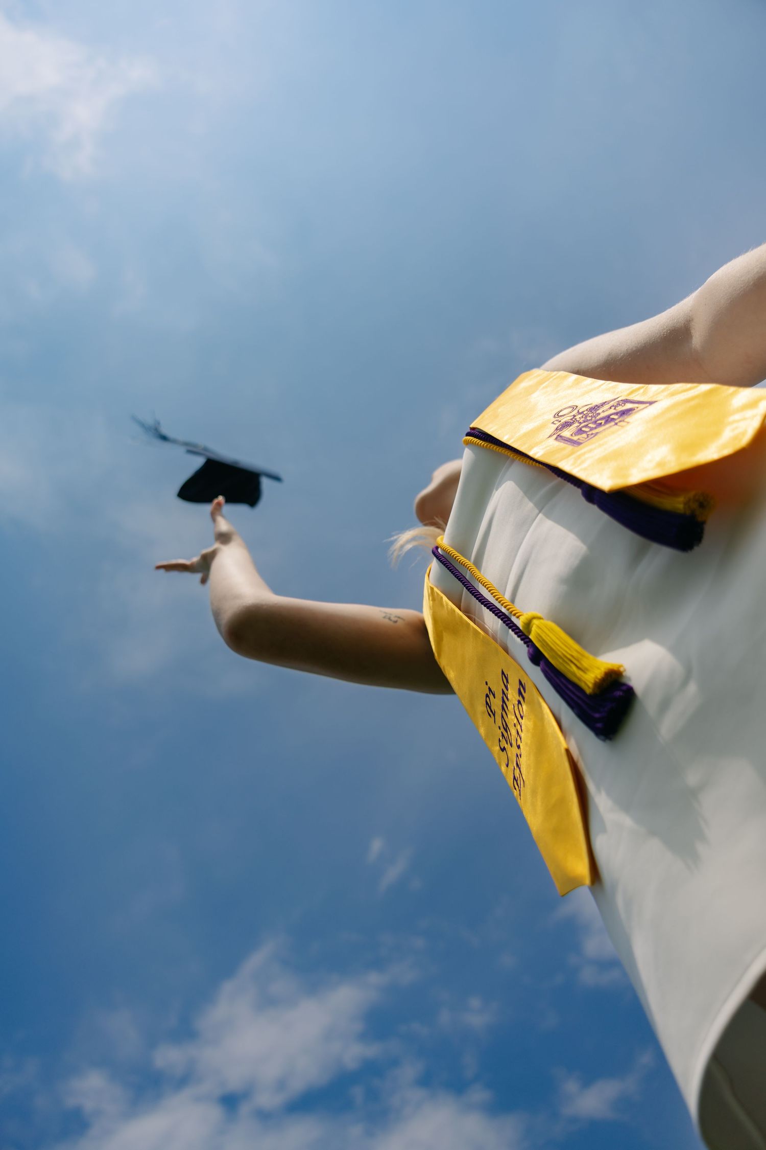 Graduate in yellow gown tosses mortarboard cap into blue sky, celebrating academic achievement outdoors.